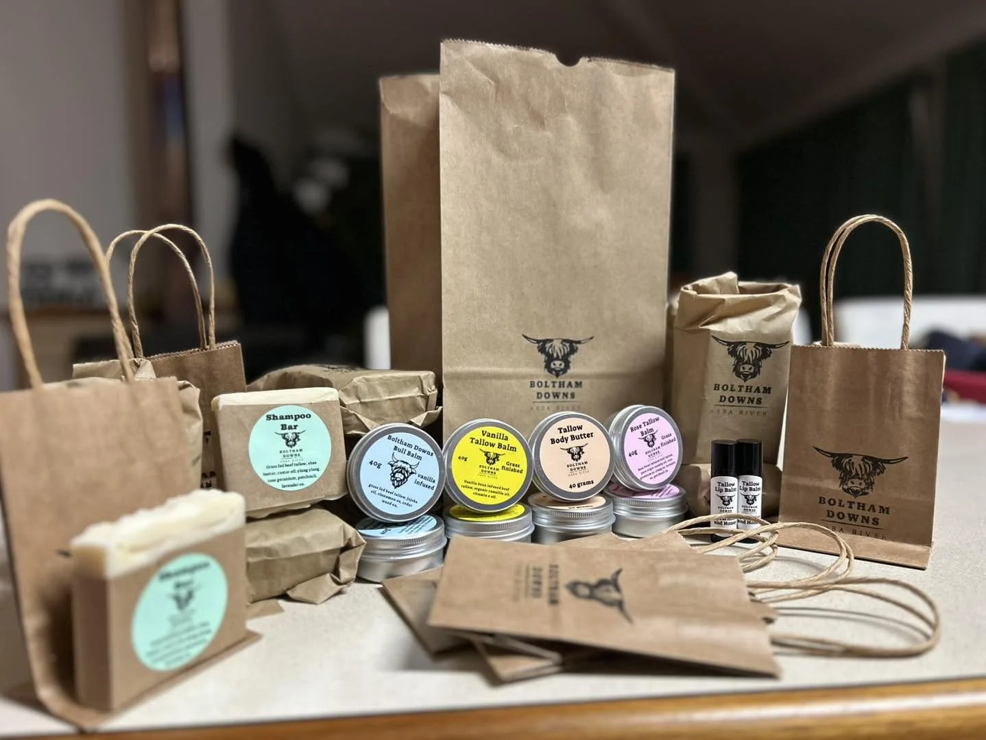 Various personal care products, including Tallow Balm, Body Butter, & Lip Balm, arranged on a table with BOLTHAM DOWNS branded paper bags and small paper gift bags with handles.