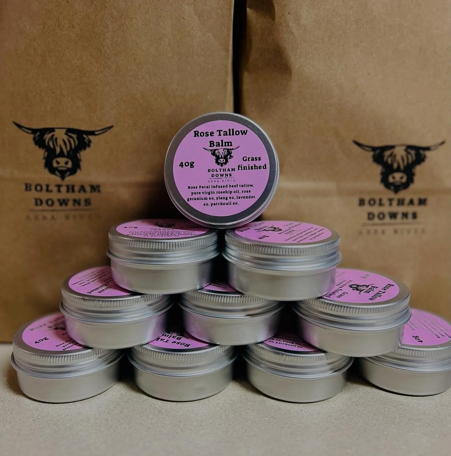 Small metal tins of Rose Tallow Balm with pink labels, stacked in a pyramid shape in front of cardboard boxes with 'Boltham Downs' logo featuring a highland cow head.
