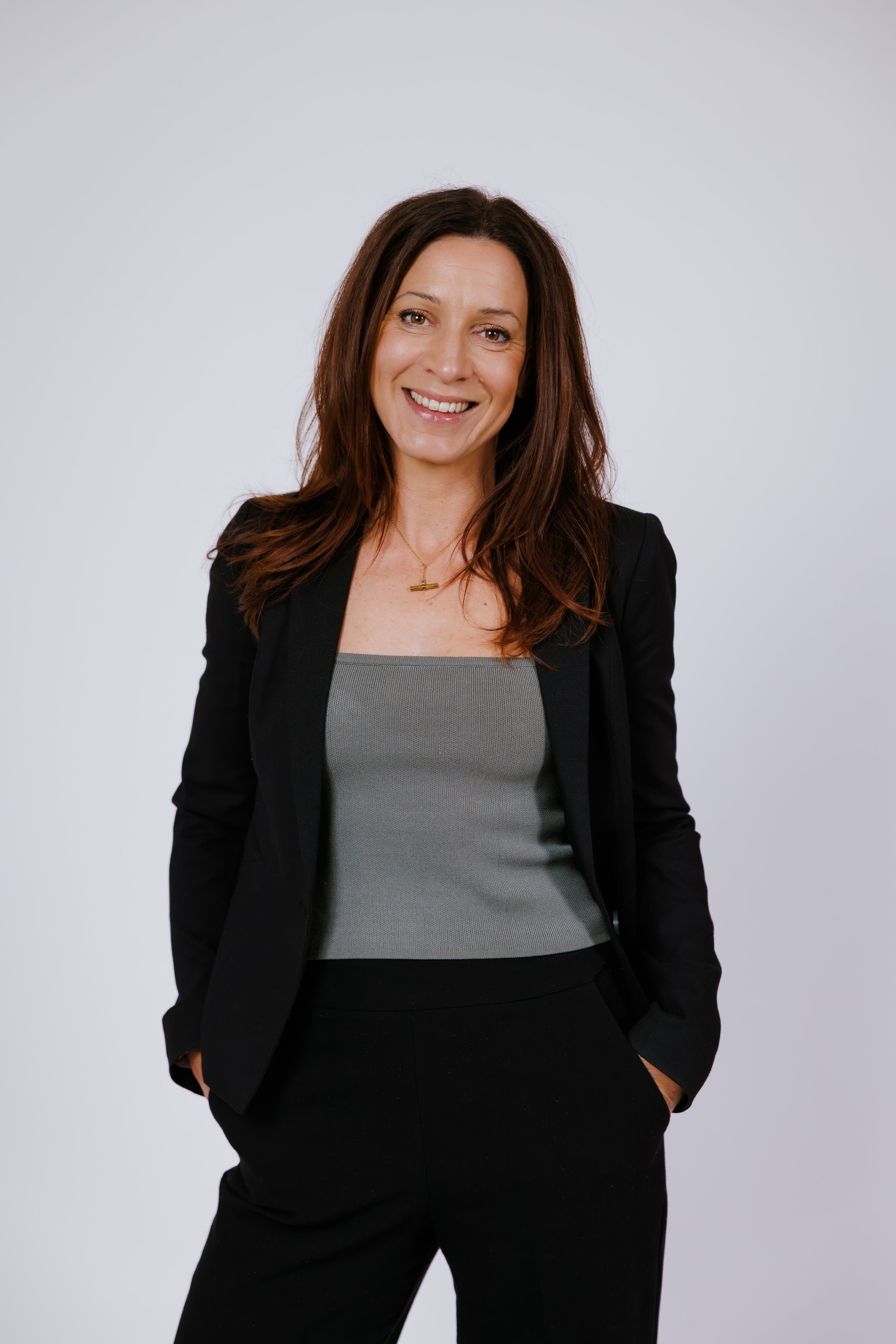 A picture of Nicole Hauxwell. She has long brown hair, is smiling, wearing a black blazer, gray top, and black pants, and is posing against a plain white background.