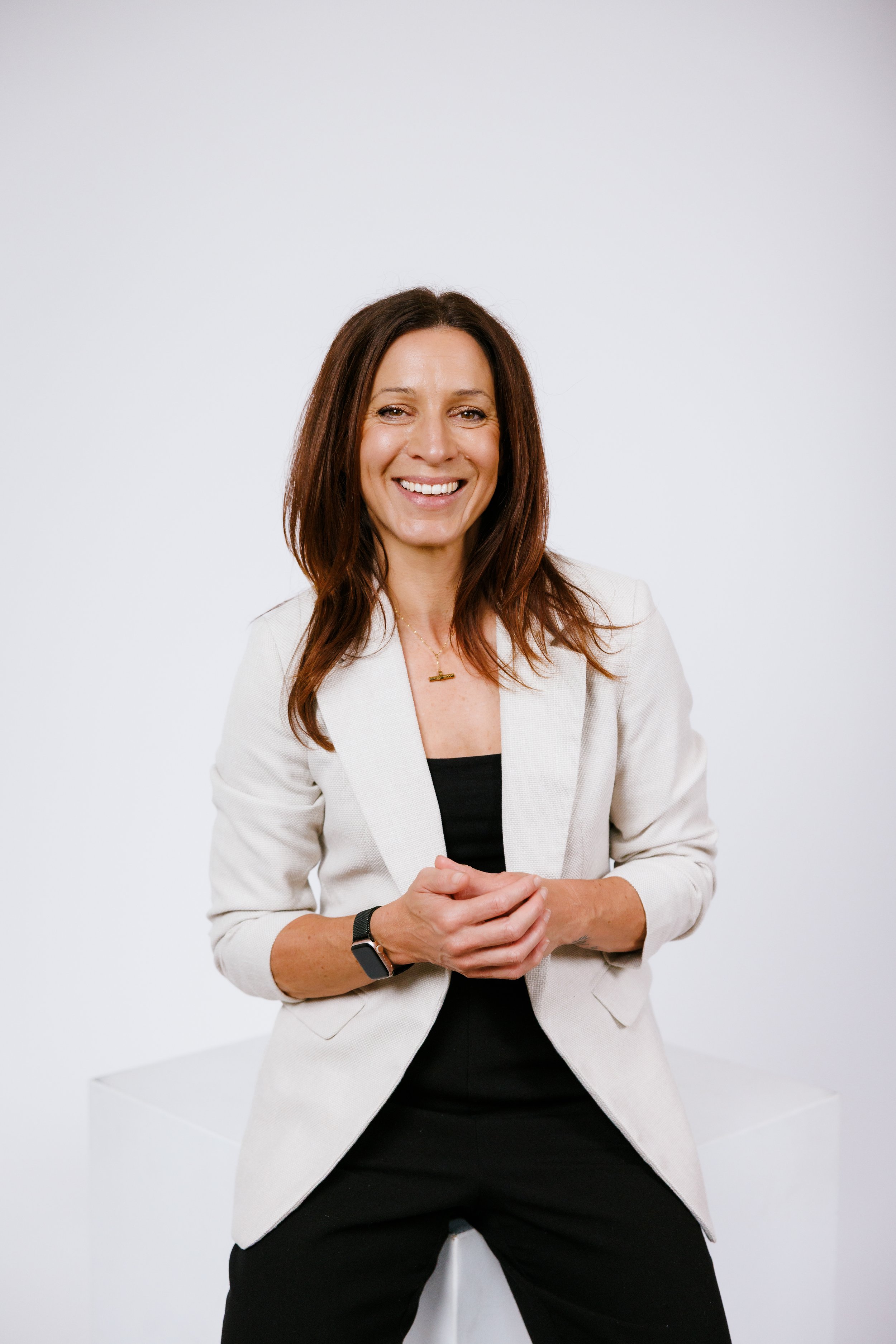A picture of Nicole Hauxwell. She has brown hair, wearing a white blazer, black top, black pants, a gold necklace, and a smartwatch, smiling and standing against a plain white background.
