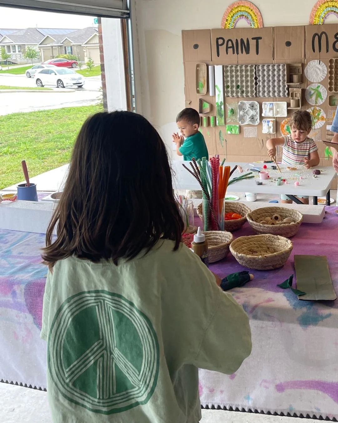 Preschooler exploring a cardboard paint wall next to a tinker table set with wooden beads, pipe cleaners, and recycled loose parts