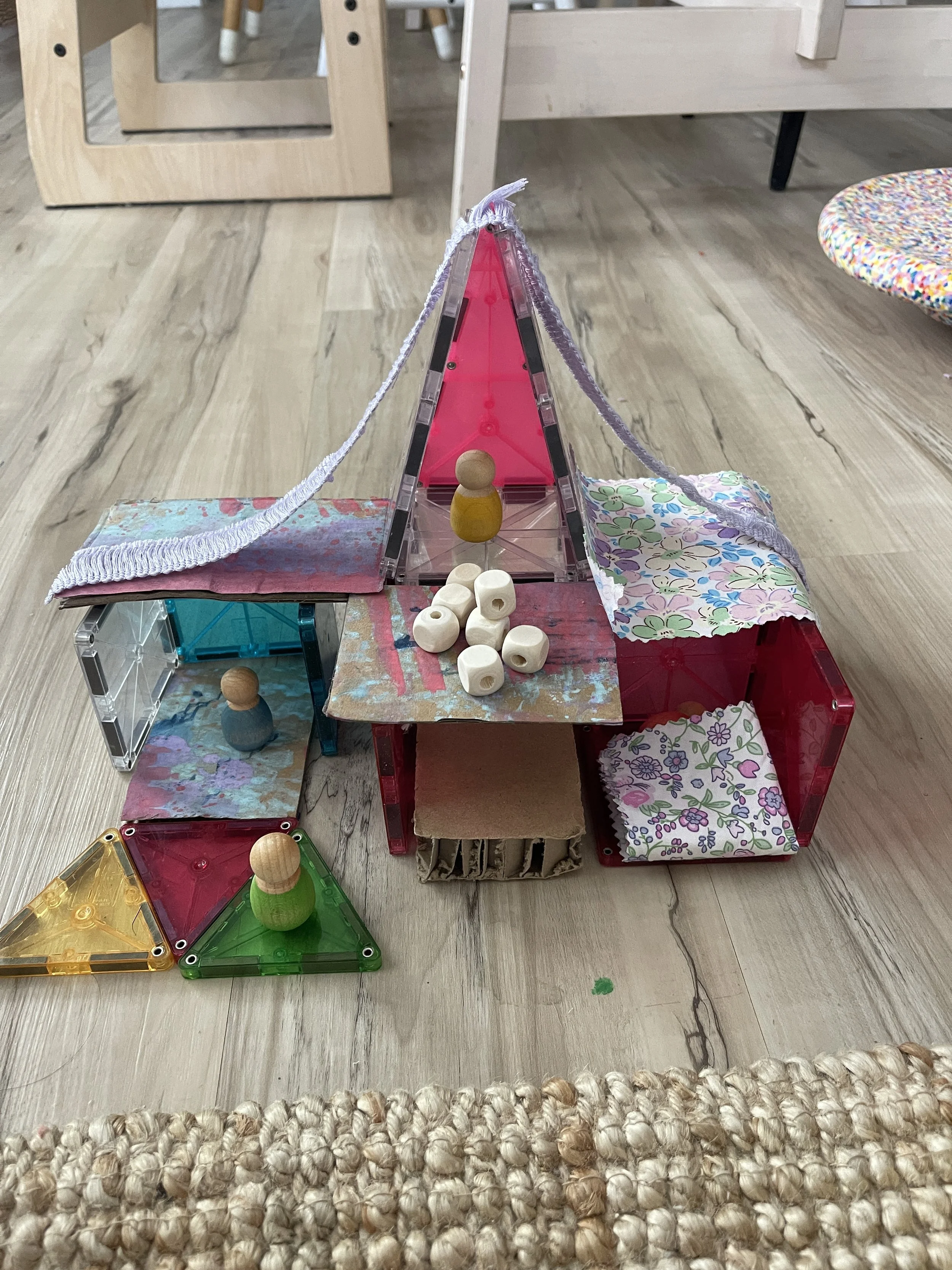 A small fort built from magnetic tiles and cardboard, draped with fabric scraps, with peg dolls placed inside as the fort's inhabitants