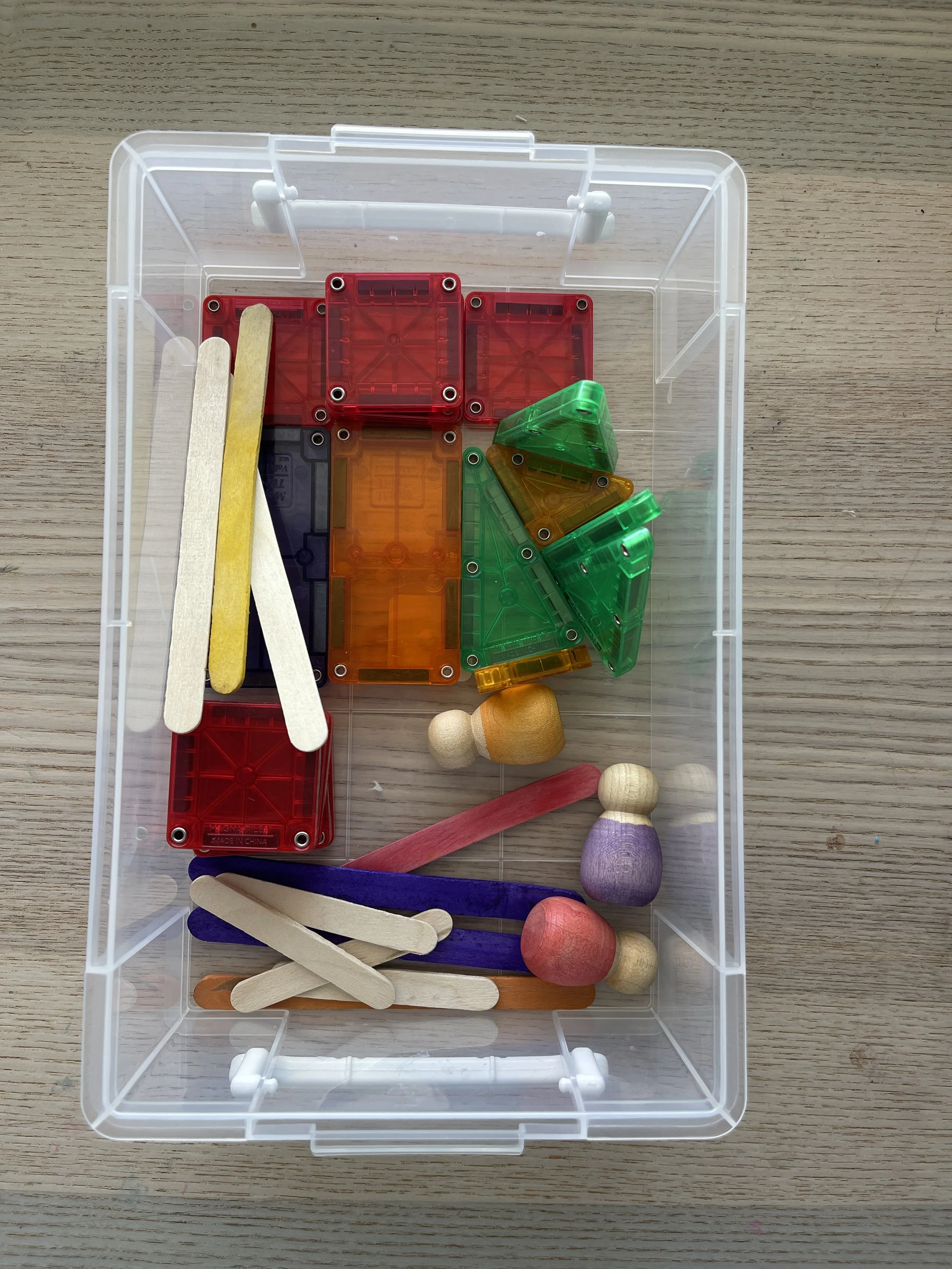 Clear bin of loose parts including magnetic tiles, popsicle sticks, and wooden peg dolls, used to build a small playground for open-ended play