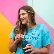 Young woman with long hair holding a drink, smiling, wearing a blue T-shirt with text, standing against a colorful geometric background.