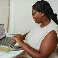 Woman with braided hair and glasses working on a laptop in an office.