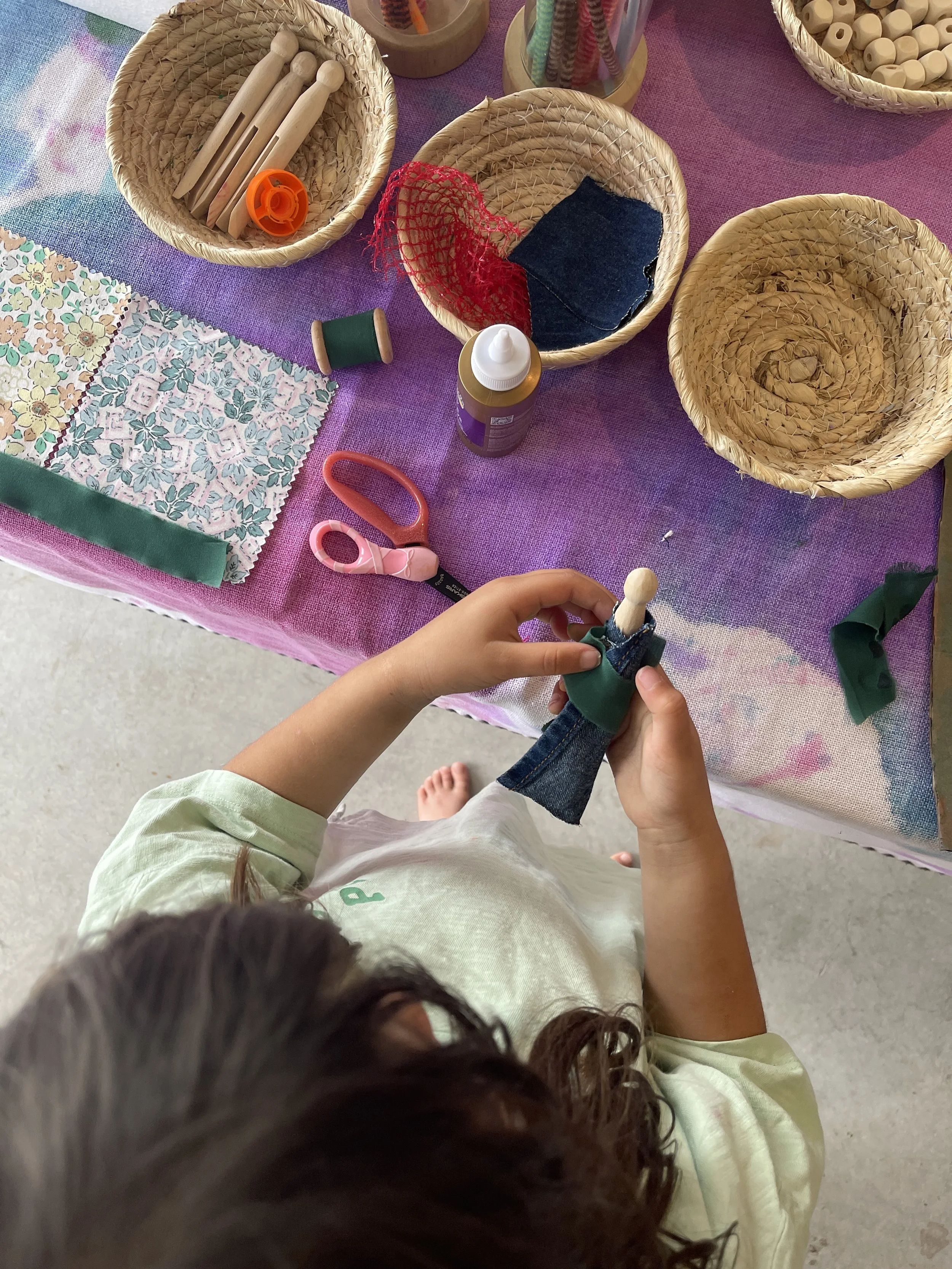 Preschooler wrapping fabric scraps around a wooden peg doll at an open-ended tinker table activity