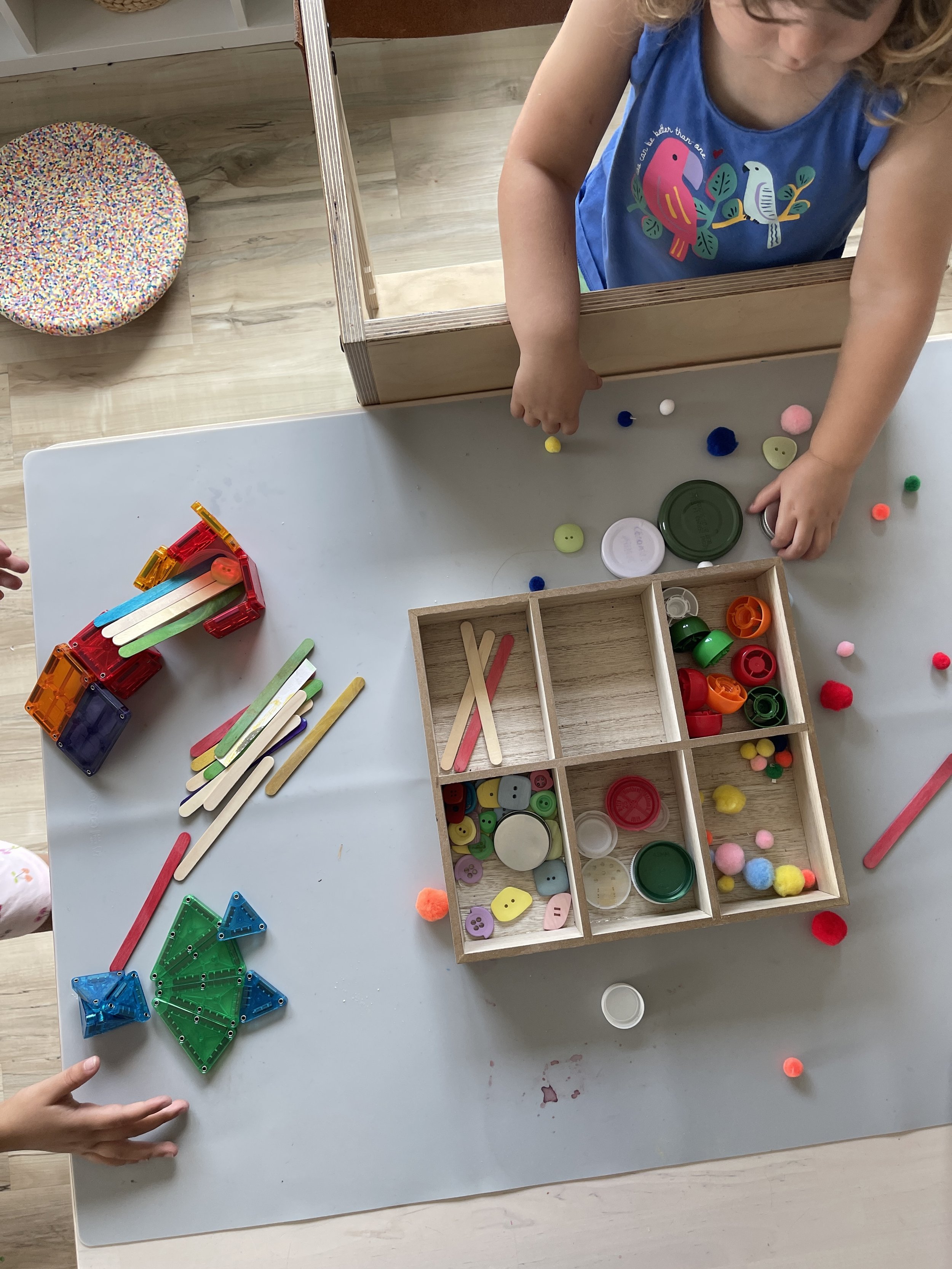 A child arranging magnetic tiles, popsicle sticks, buttons, and pom poms from a wooden compartment tray into a loose parts scene