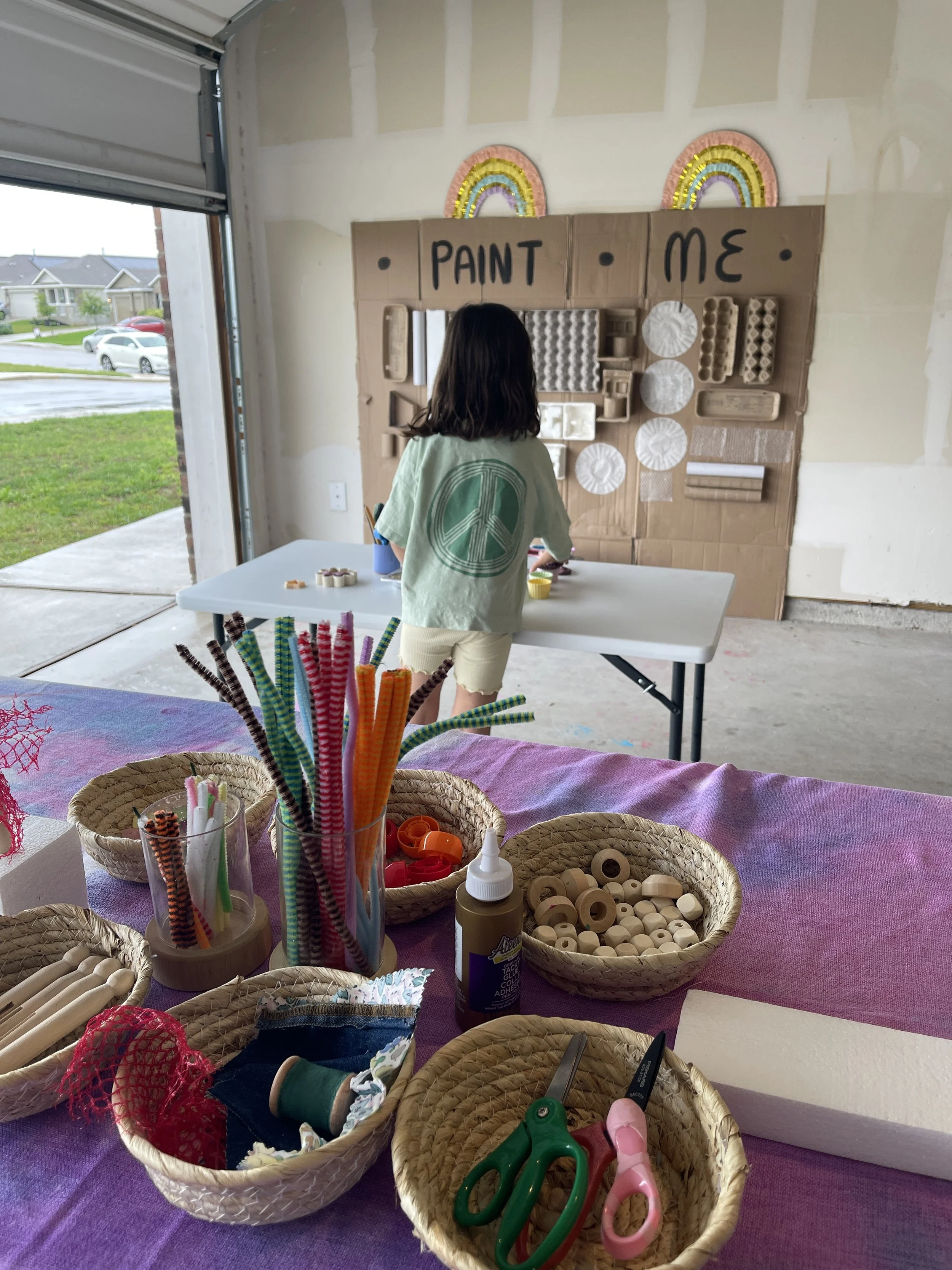 Preschooler exploring a cardboard paint wall next to a tinker table set with wooden beads, pipe cleaners, and recycled loose parts