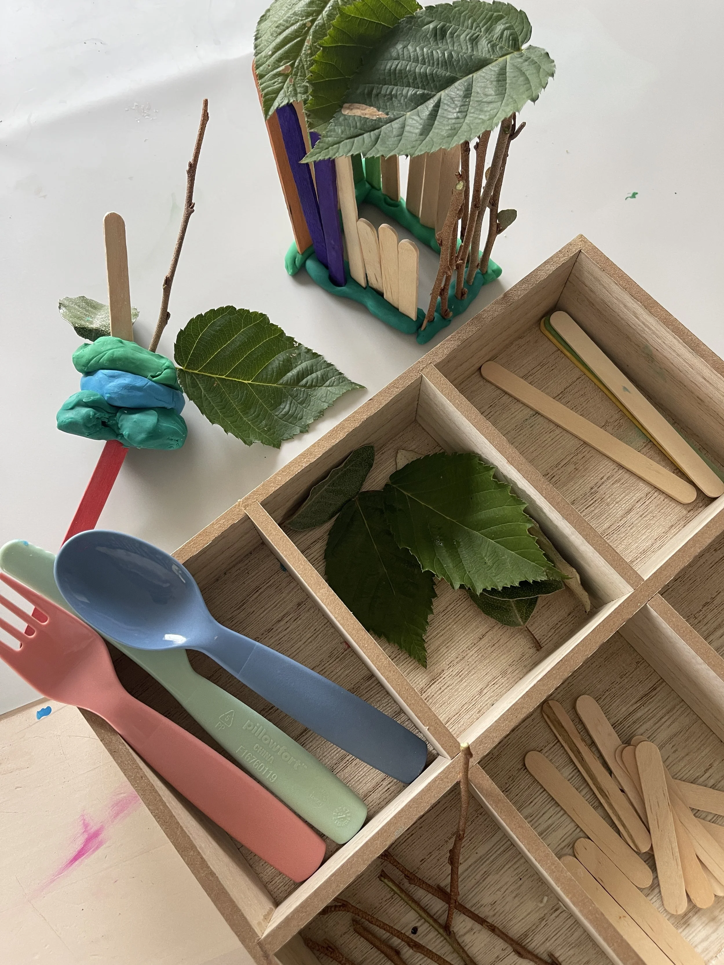 Twigs, popsicle sticks, and leaves pressed into a clay base to build a small nature structure, arranged in a wooden compartment tray