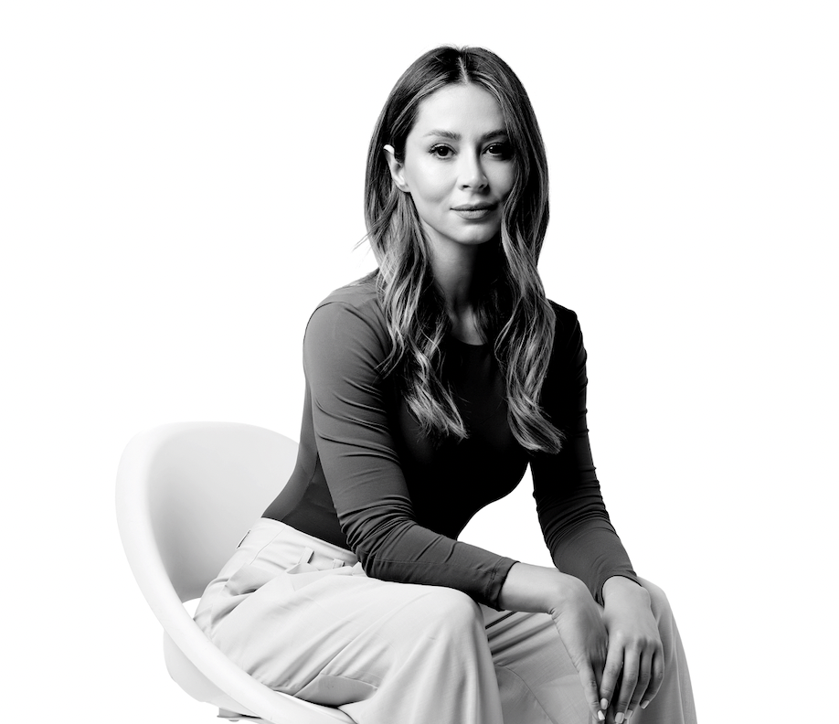 Black and white portrait of a woman sitting on a modern chair, wearing a long-sleeve top and light-colored pants, looking at the camera with a calm expression.