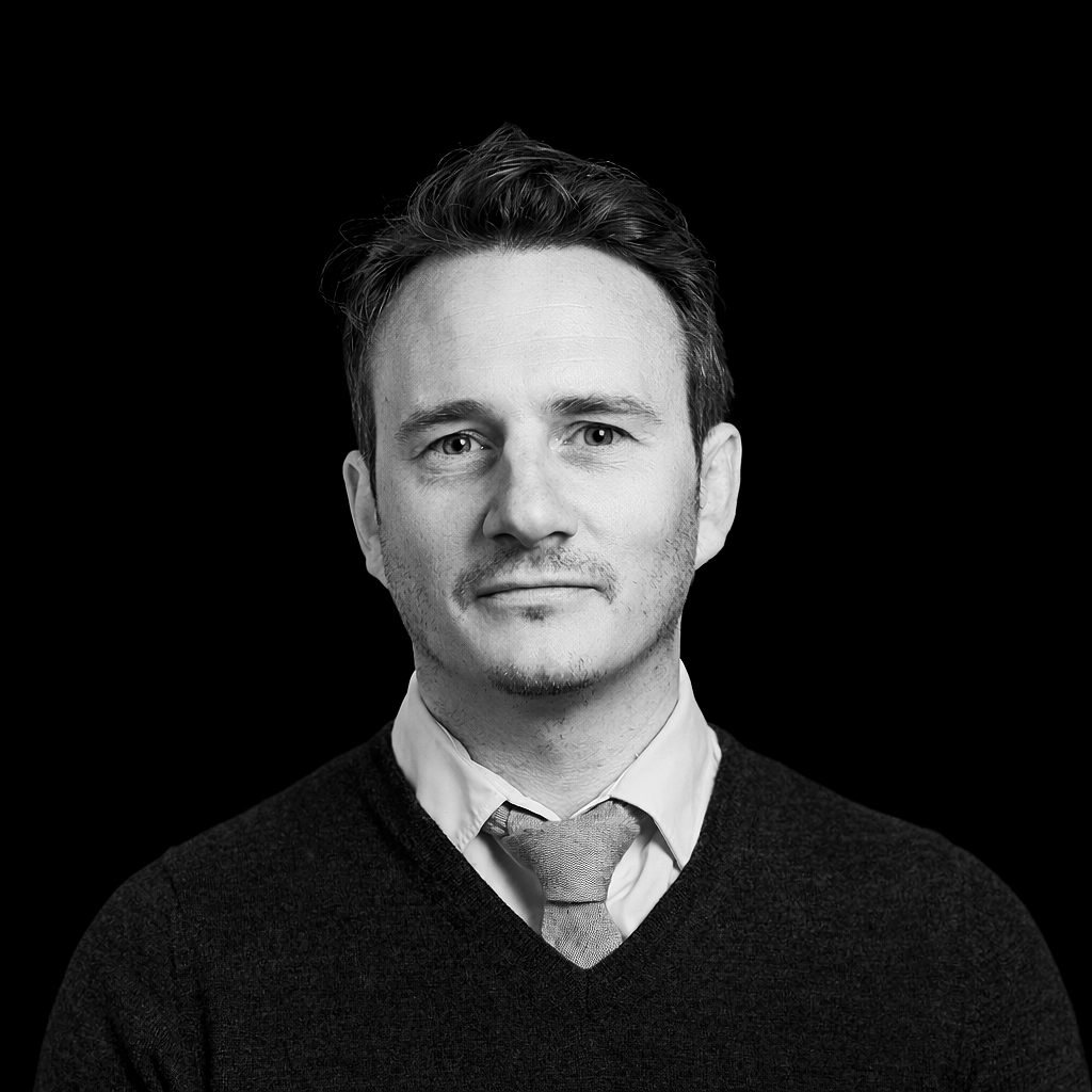 Black and white portrait of a man with short hair, wearing a collared shirt, tie, and sweater, against a dark background.