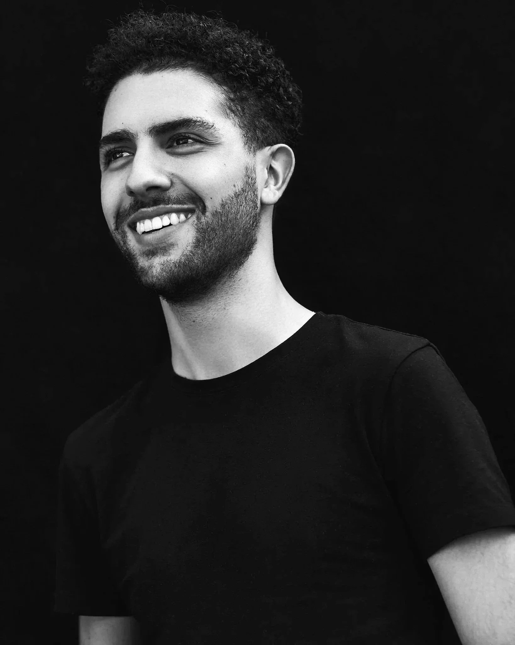 Black and white close-up photo of a man with curly hair, a beard, smiling and looking to his left, wearing a black shirt against a solid dark background.