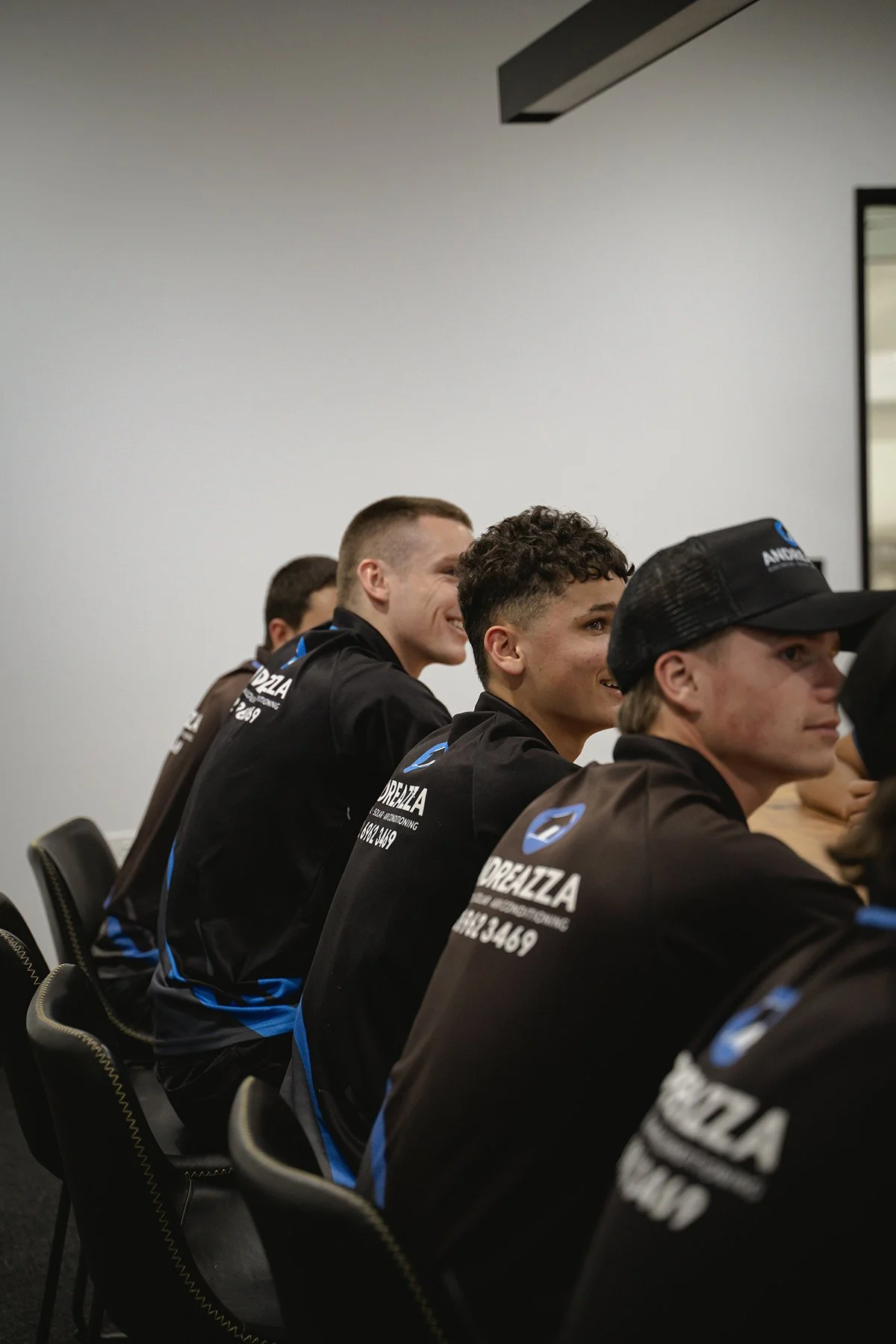 A group of young men in black and blue sports jackets sitting at a conference table, smiling and listening attentively.