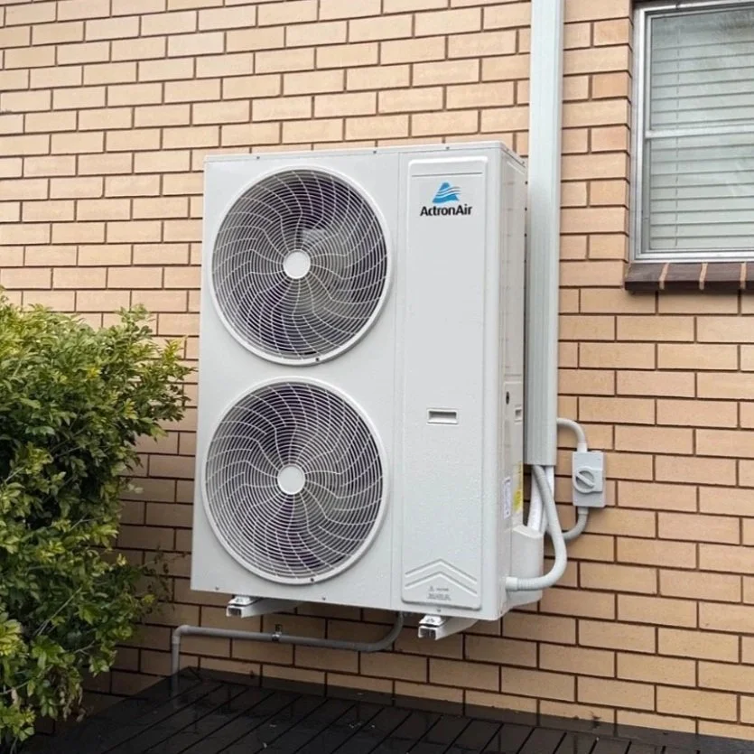 A white air conditioning or heat pump outdoor unit is installed on the exterior brick wall of a building. It features two large circular fans.