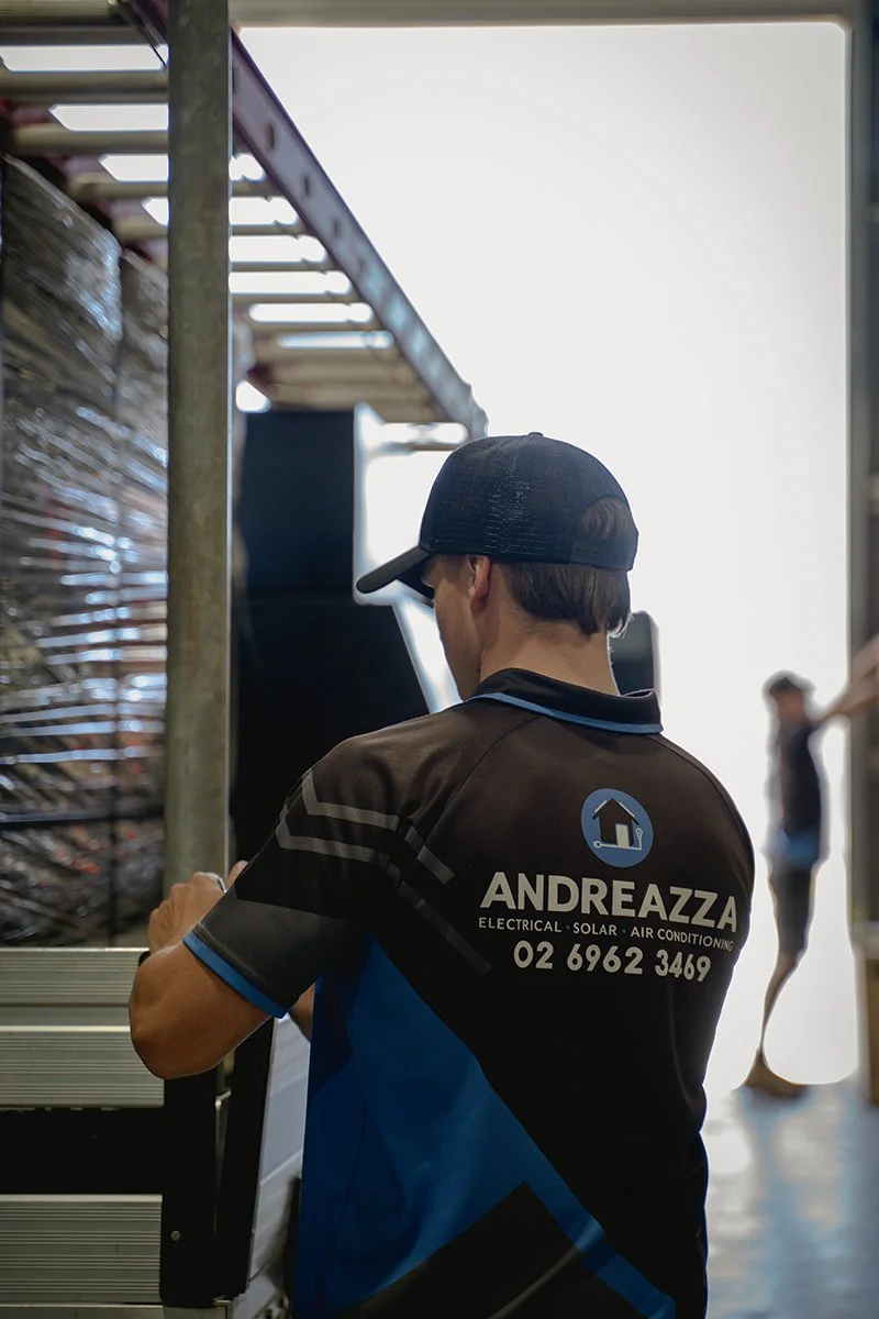 A man wearing a black cap and a black and blue shirt with the text 'ANDREAZZA ELECTRICAL SOLAR AIR CONDITIONING' and a phone number, standing in a workplace next to equipment, with a woman in the background.