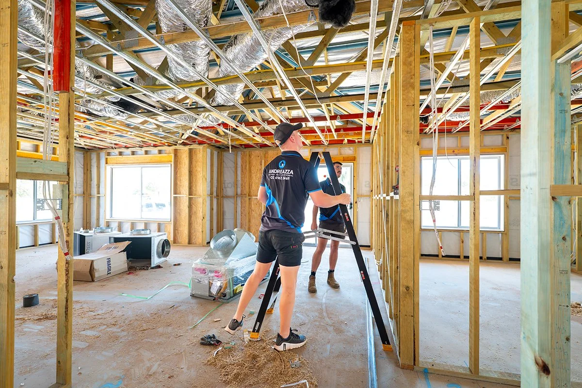 Two workers in black and blue uniforms working inside a house under construction, with wooden framing, windows, and exposed ceiling ductwork.