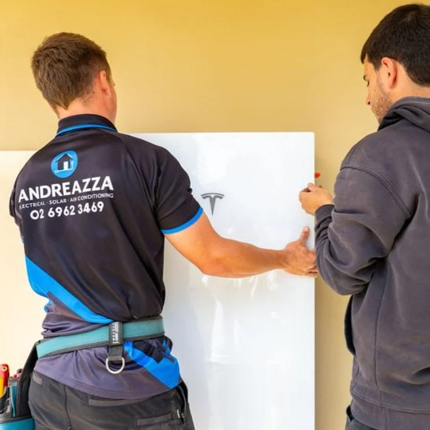 Two men are working on a Tesla vehicle, with one man wearing a shirt labeled 'ANDREAZZA Electrical, Solar, Air Conditioning' and the other man holding a marker, both discussing or analyzing something related to the car.