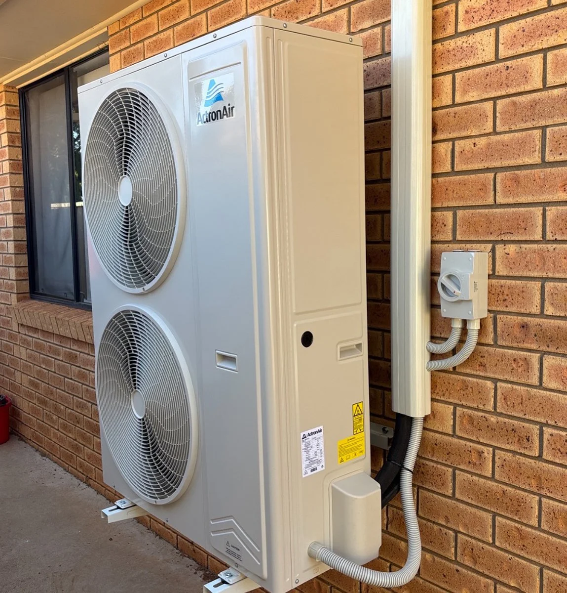 An outdoor air conditioning unit with two large fans mounted on a beige metal cabinet, installed on a brick wall outside a house, with a window nearby.