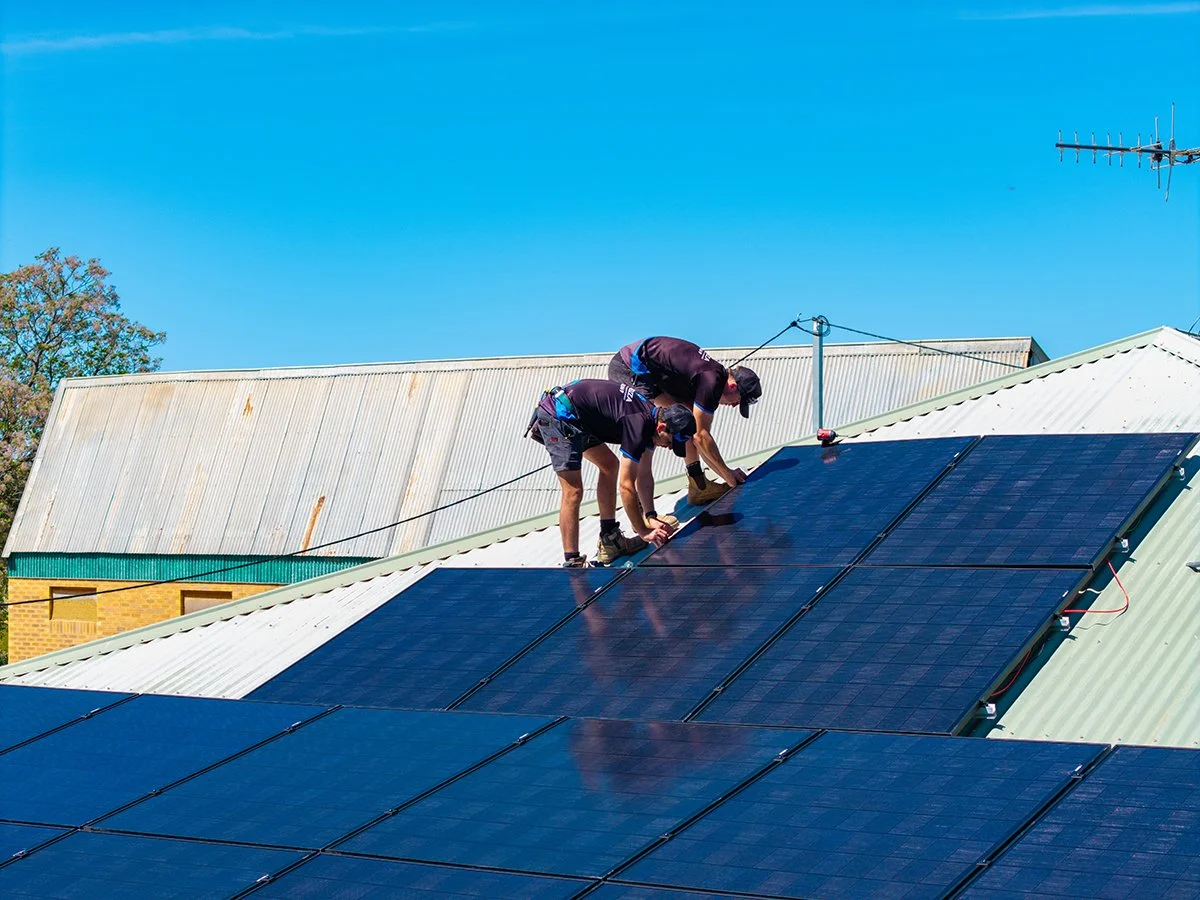 Two workers installing solar panels on a sloped roof, with a clear blue sky in the background.