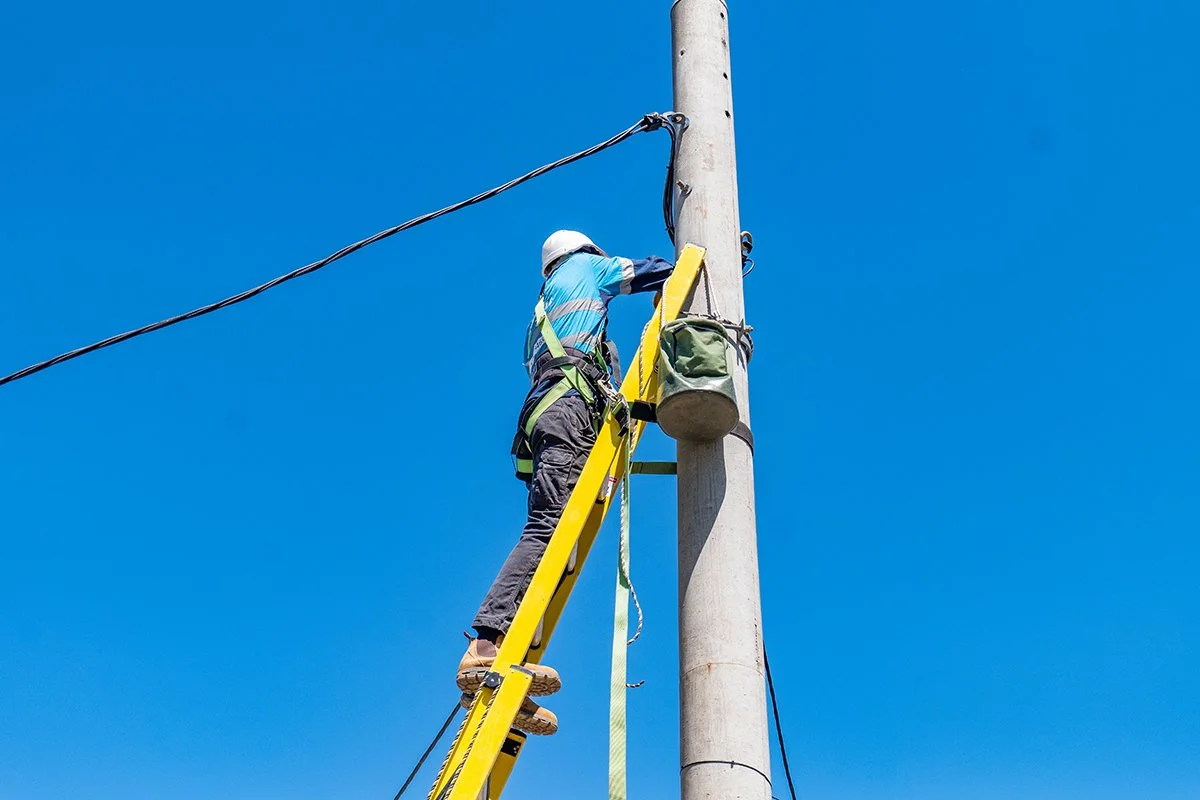 A utility worker in safety gear working on power lines attached to a tall utility pole against a clear blue sky.