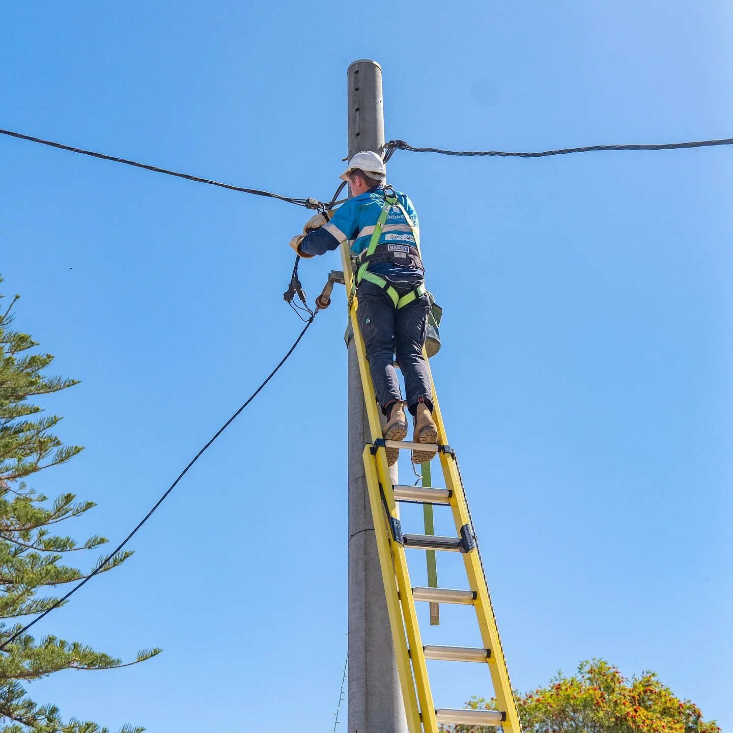 A worker in safety gear on a ladder repairing power lines on an electrical pole against a clear blue sky.
