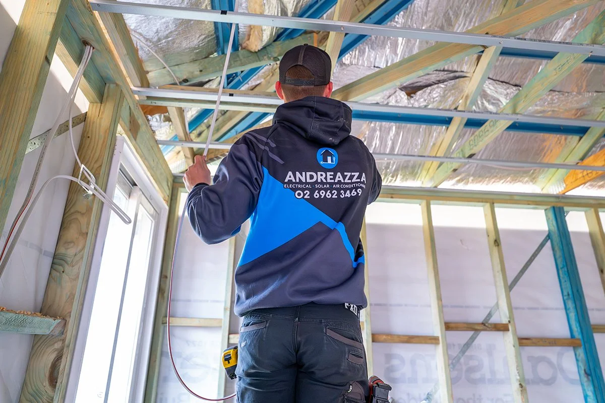 A man working on electrical wiring inside a building under construction, wearing a hoodie with a company logo and contact information.
