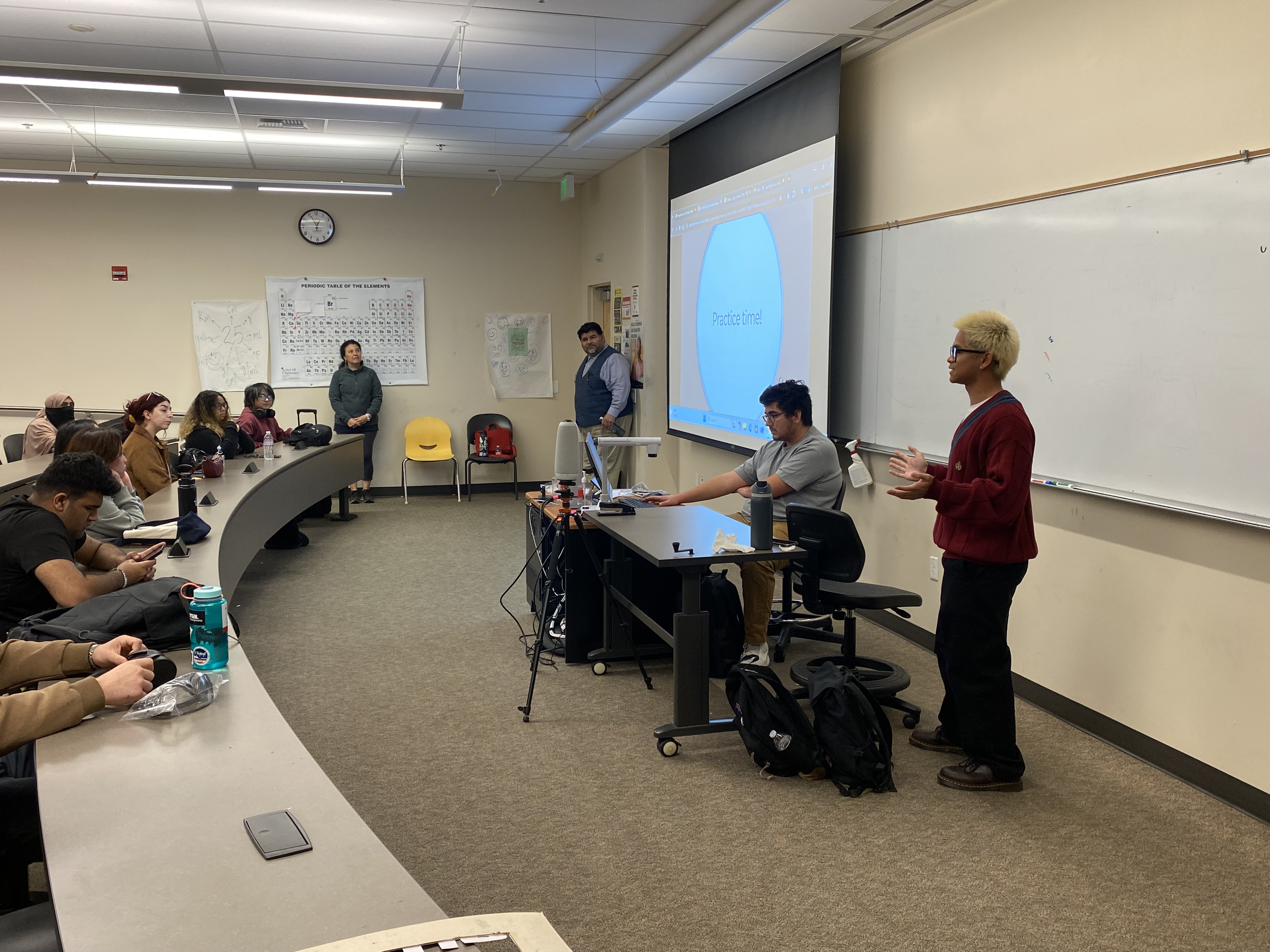 A classroom with students and two instructors. One instructor is standing and speaking, while the other is seated at a desk with a computer. Students are seated facing the front, some taking notes or using their phones. A large screen displays a slid
