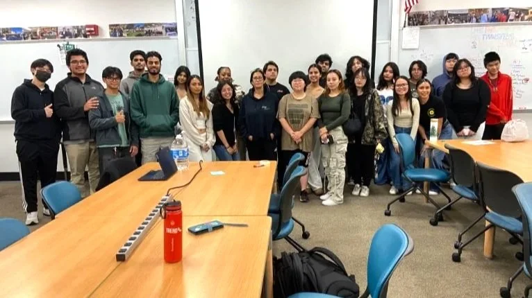 Group of about 18 people standing in a classroom or meeting room, facing the camera, with tables and chairs in the foreground.