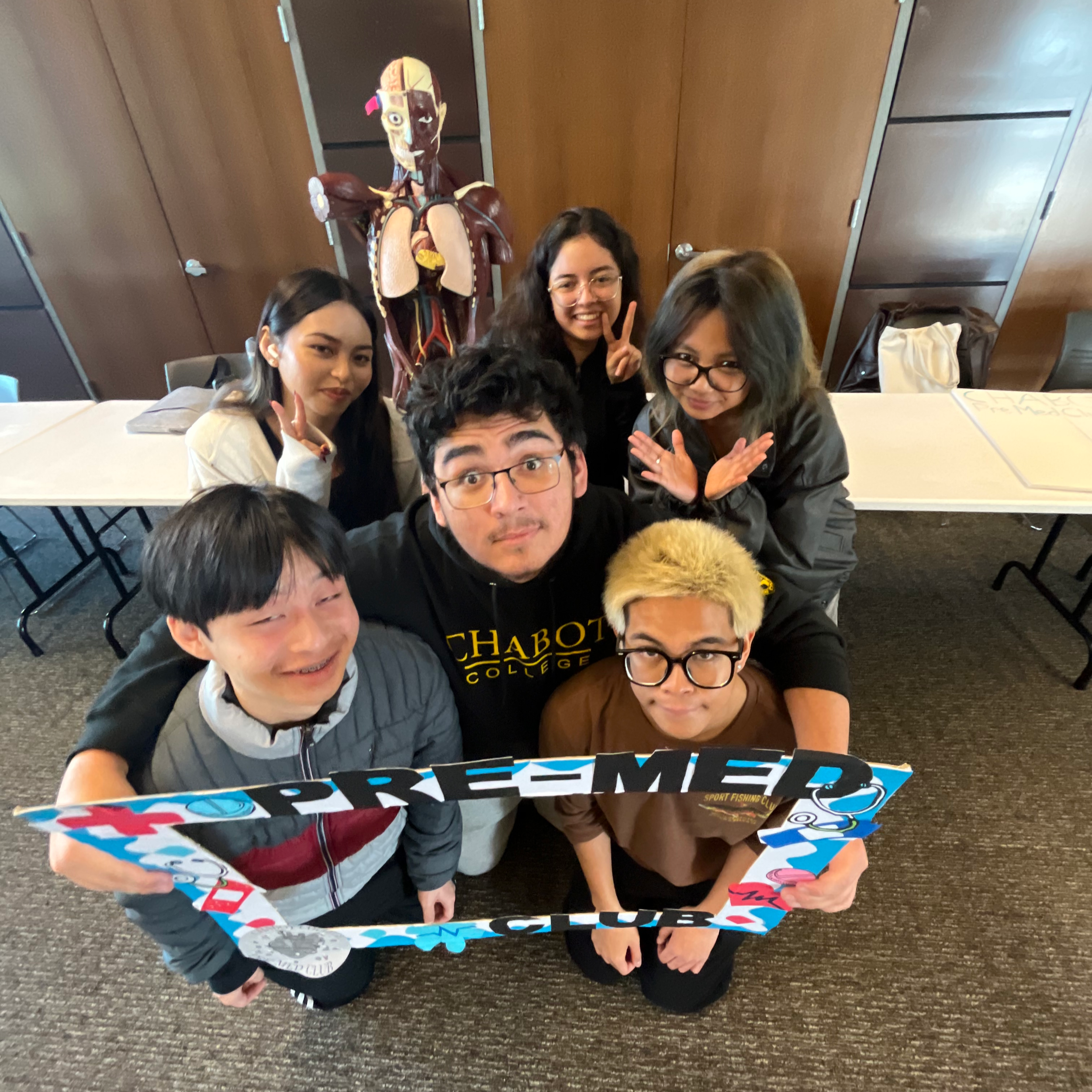 Group of seven young people smiling and posing with a colorful border frame, standing in a room with a life-sized anatomy model in the background.