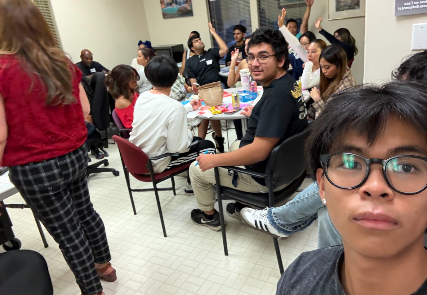 A group of diverse people gathered in a room around tables, some raising their hands, with one person taking a selfie in the foreground.