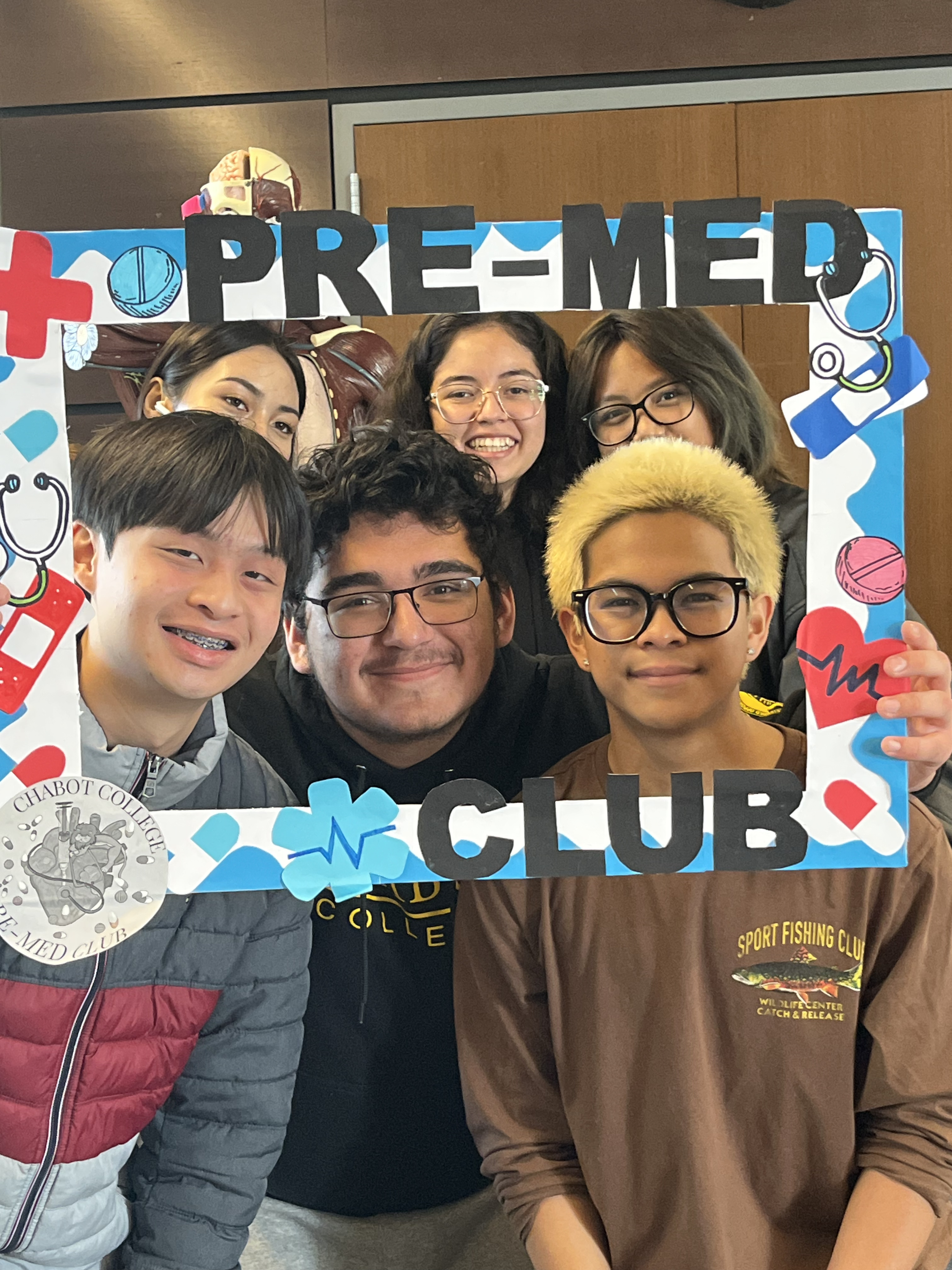 Group of six young people smiling inside a decorated frame that reads 'Pre-Med Club', with medical-themed drawings like stethoscopes, hearts, and pills, at an indoor event.