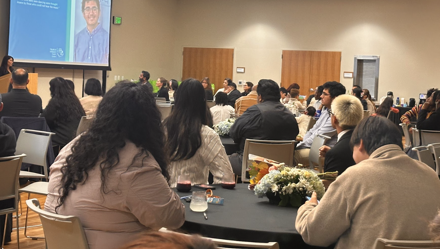 Conference room filled with attendees seated at round tables, listening to a speaker in front of a large screen displaying a man in glasses and a blue shirt.
