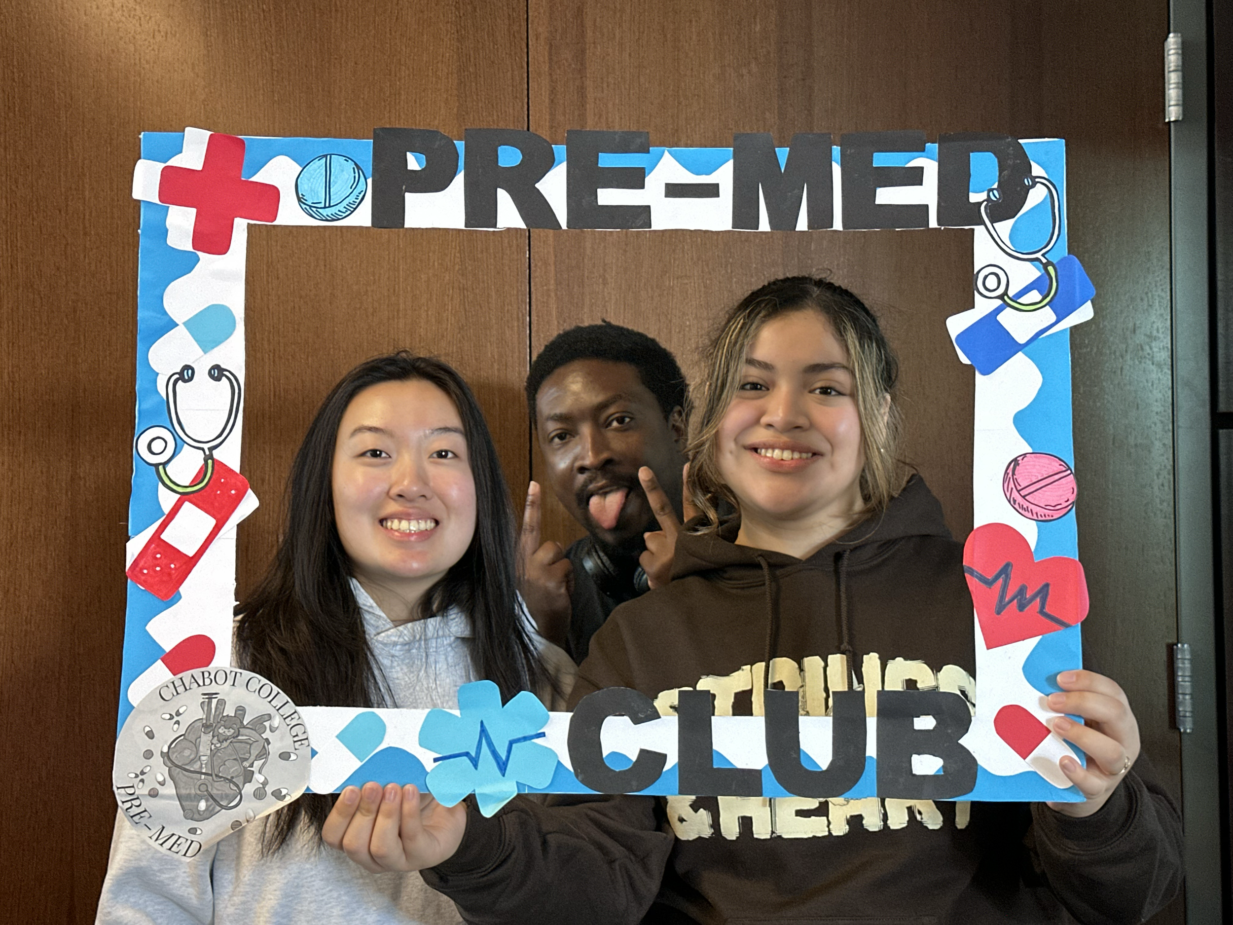 Three smiling people holding a framed sign that says "PRE-MED CLUB" with medical-themed decorations, including a stethoscope, heart, and pills.