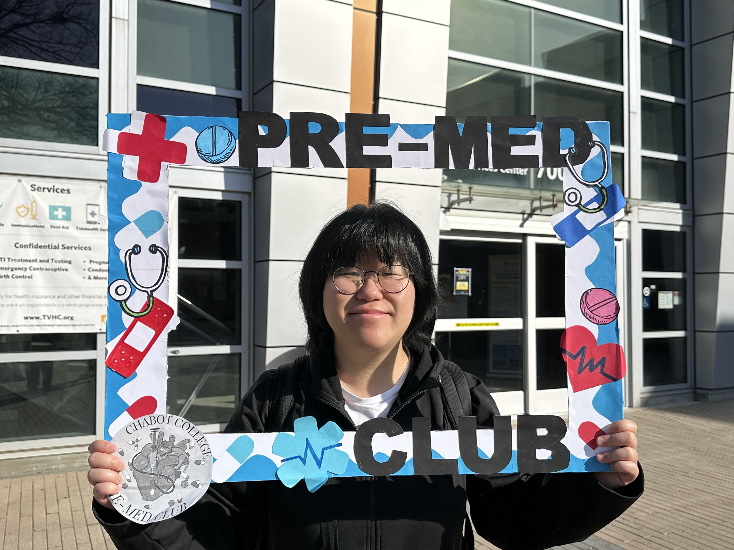 Person holding a decorated frame with the words 'Pre-Med Club' outside a building with glass windows.