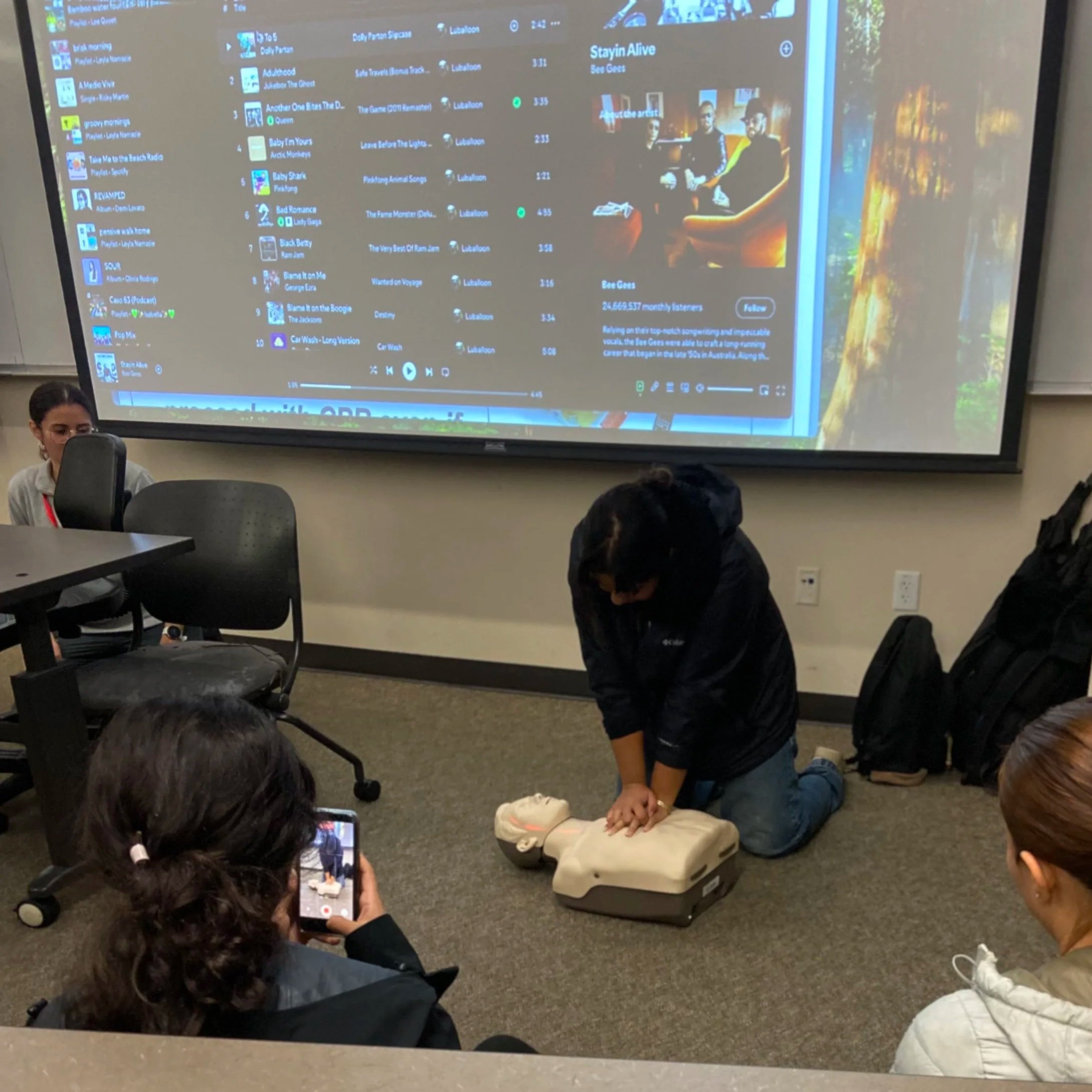 A person is performing CPR on a mannequin in a classroom setting while others observe. A large screen behind shows a computer list with music tracks and album details.
