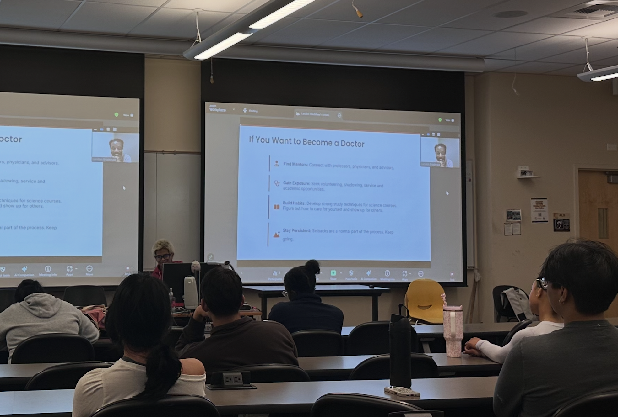 Classroom with students attending a presentation on becoming a doctor, displayed on two large screens, with a woman at the front and students seated facing the screens.