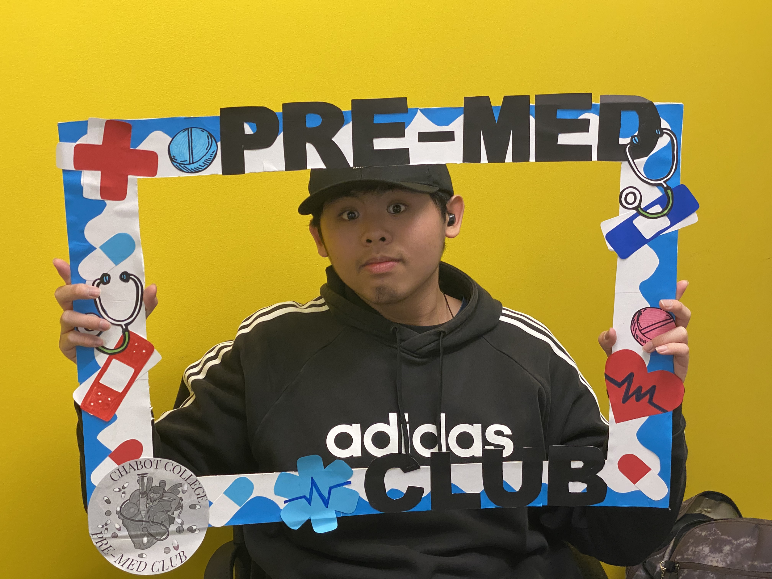 A young man wearing a black Adidas hoodie and a black cap, holding a decorated frame with medical-themed illustrations, stands against a bright yellow wall. The frame has the words 'Pre-Med Club' and features drawings of scrubs, stethoscopes, bandage