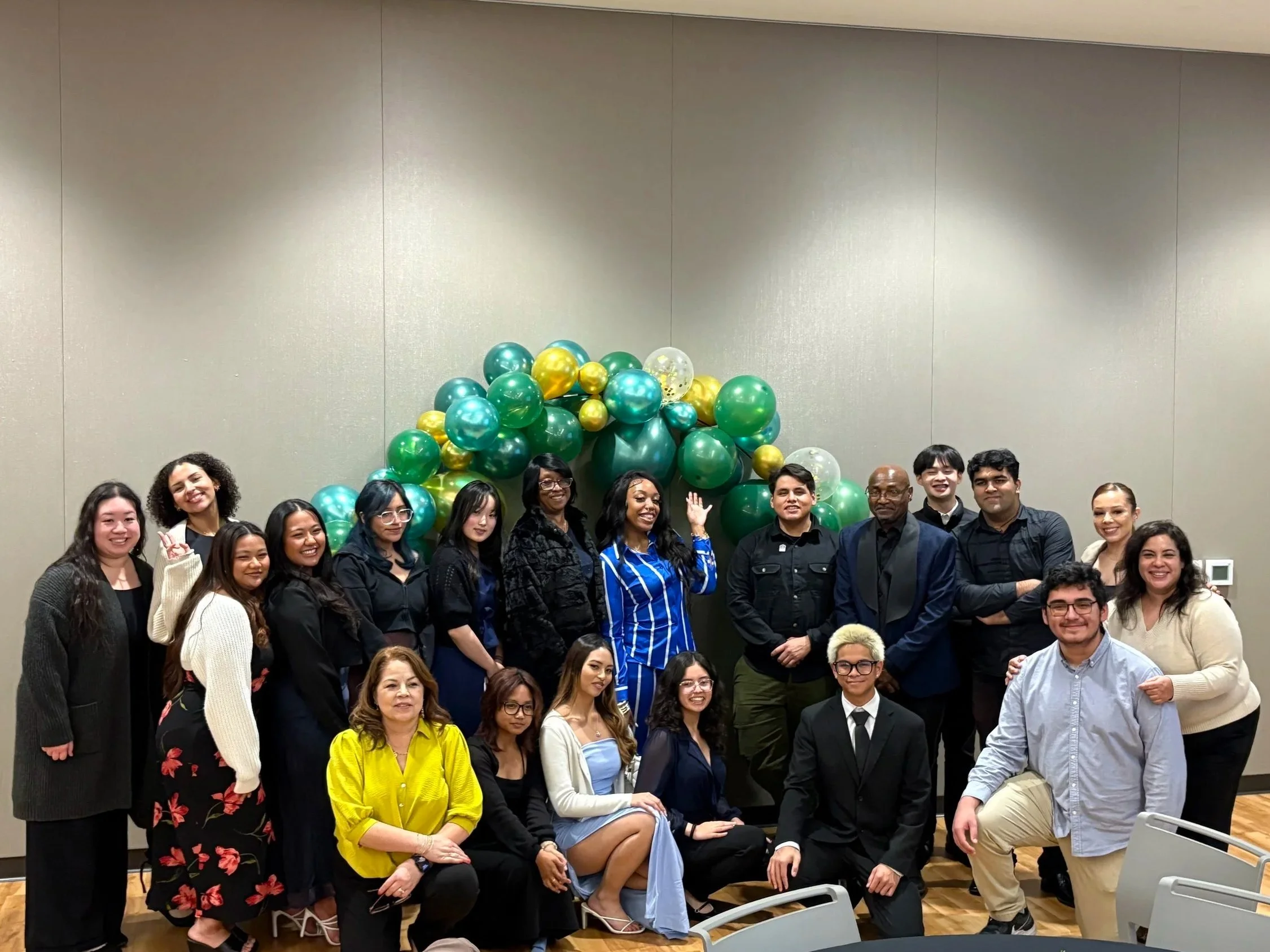 Group of 17 diverse people posing in front of green, gold, and clear balloons arranged in an arch in a conference room with a gray wall.