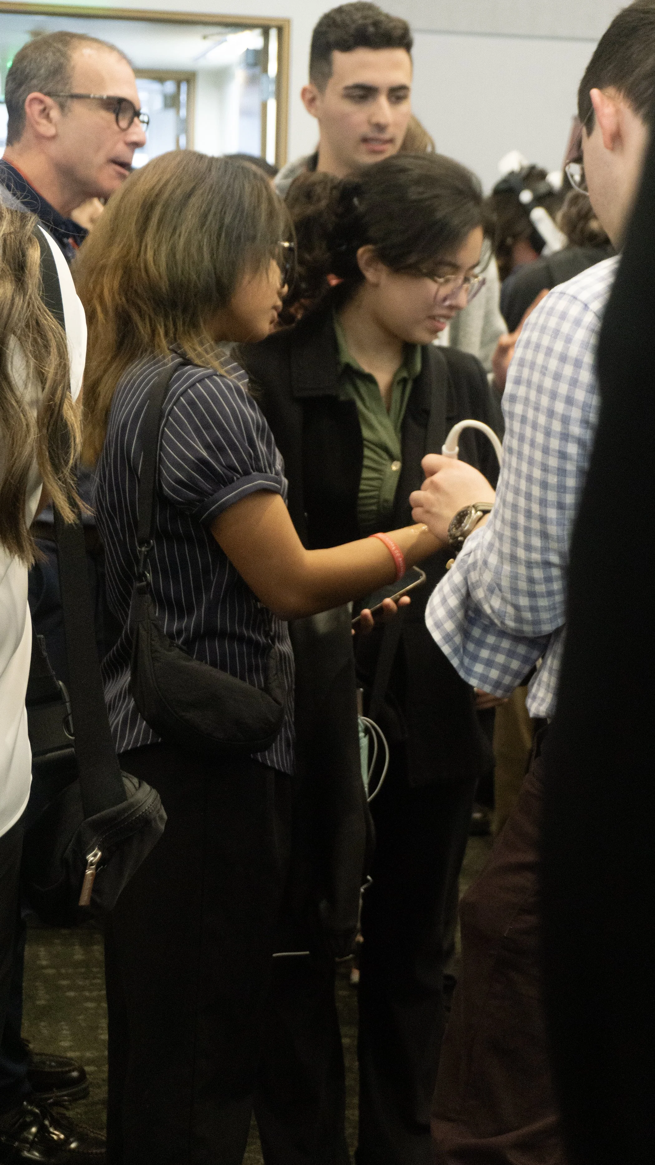 People gathered in a crowded indoor space, with a young woman in glasses and a green shirt looking at a smartphone, surrounded by several adults.