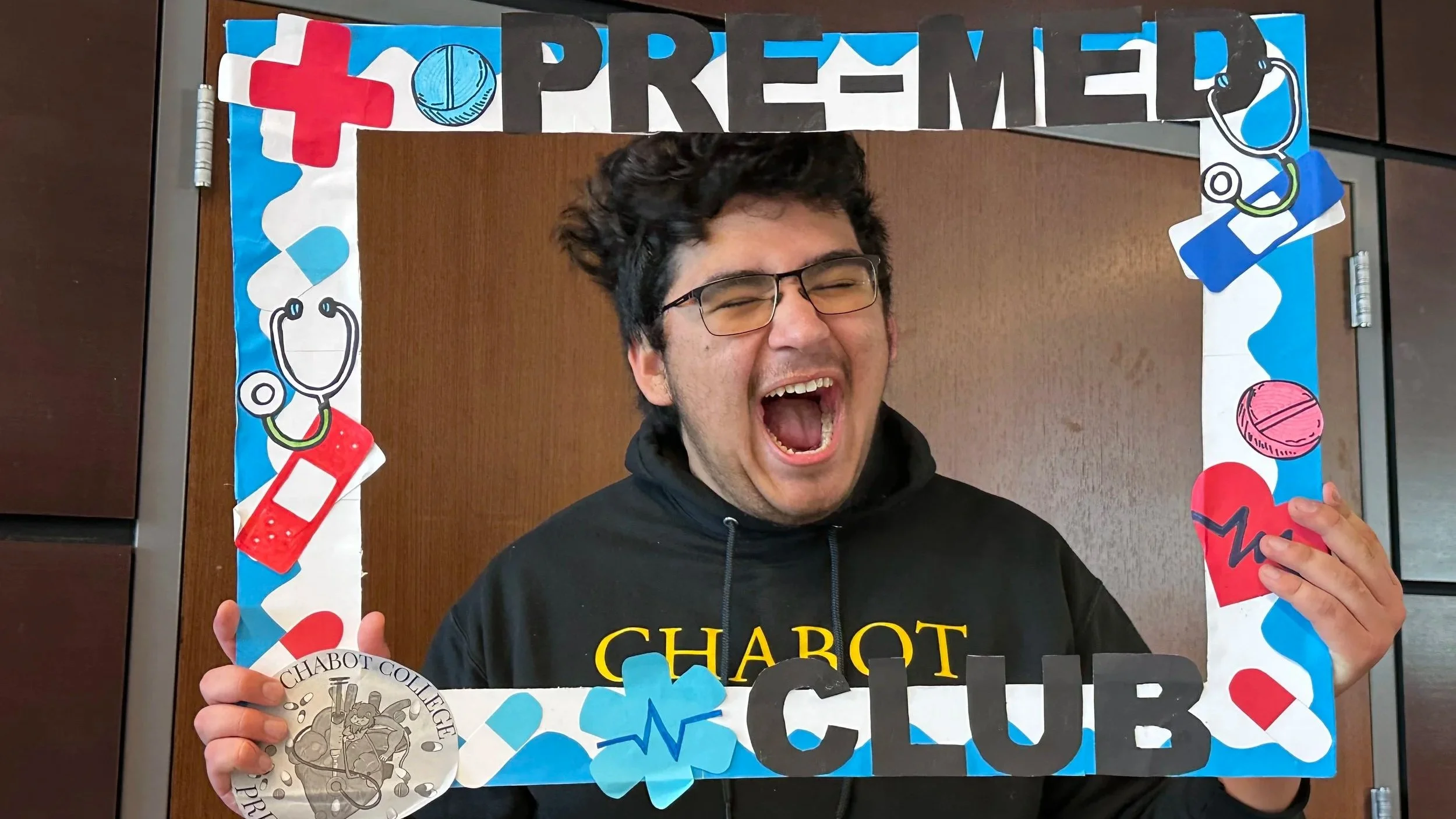 Young man wearing glasses, smiling, holding a photo frame decorated with medical-themed cutouts, including stethoscopes, pills, heart, and hearts, with the text 'PRE-MED CLUB' on top.