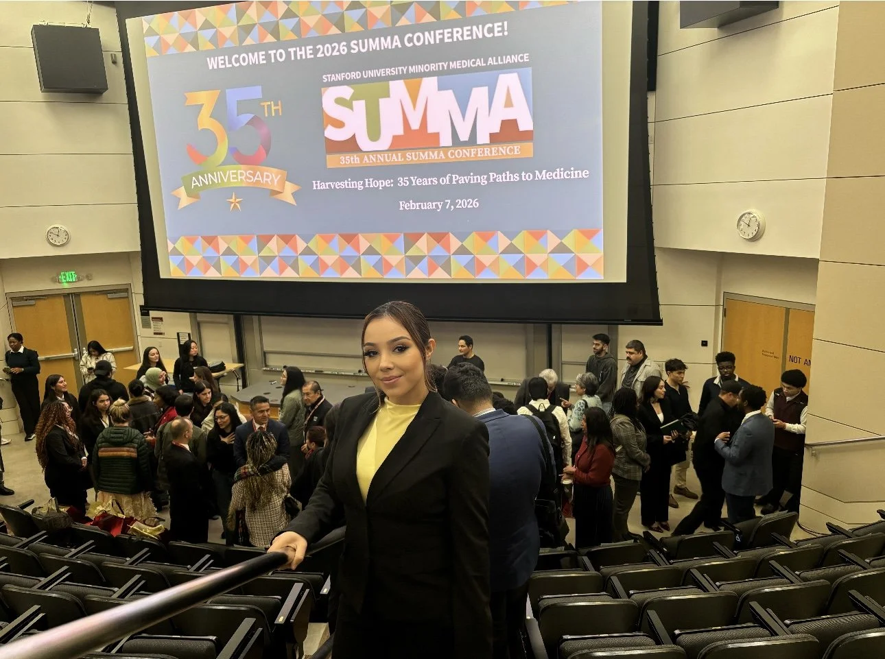 A young woman in business attire stands looking at the camera in a lecture hall filled with attendees. In the background, a large screen displays the 35th Annual Summa Conference, celebrating its 35th anniversary with the theme "Harvesting Hope: 35 Y
