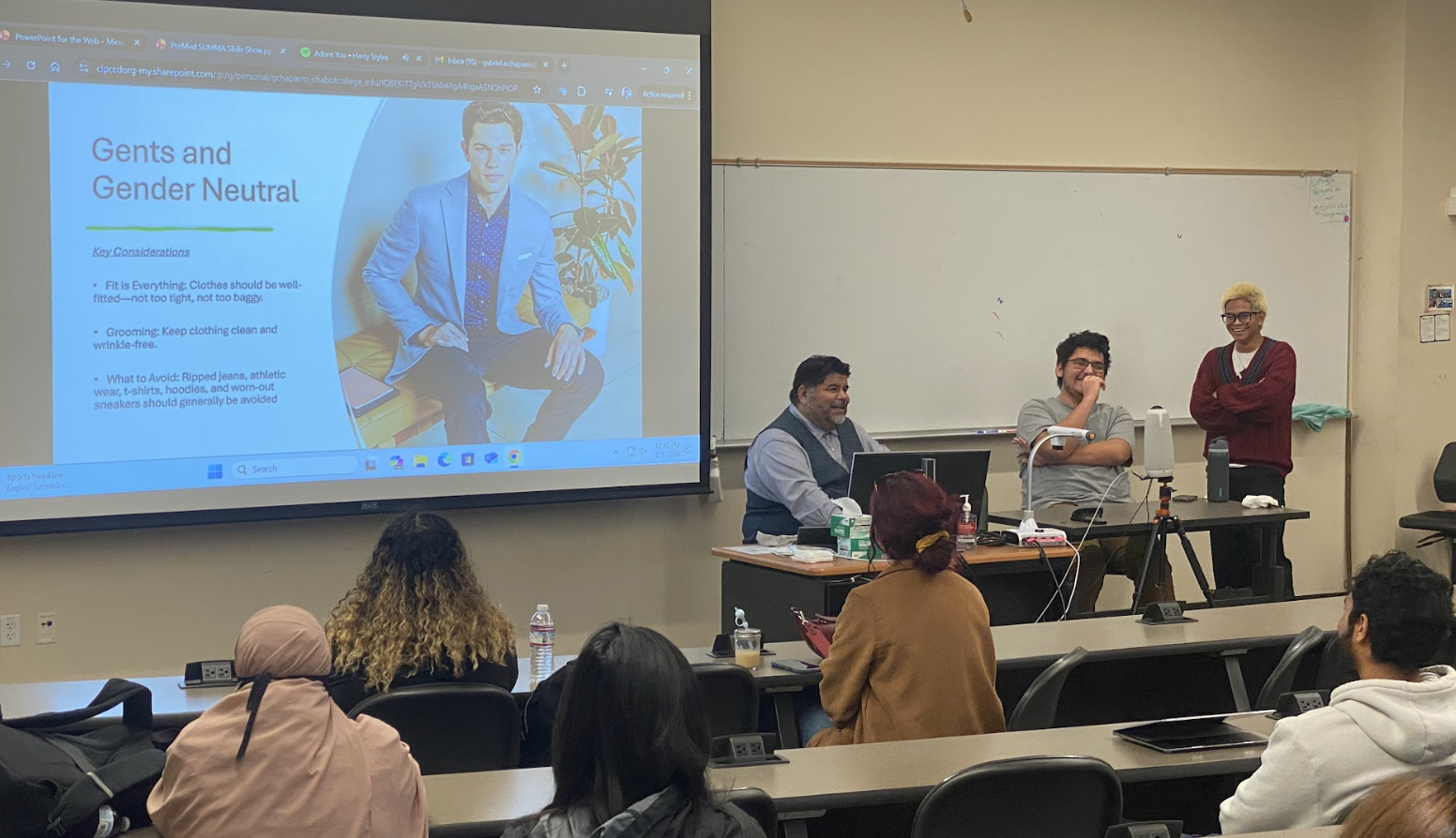 A classroom presentation on gents and gender neutral fashion with a projected slide showing a young man in a blue suit and several bullet points, three speakers at the front of the room, and students seated at desks watching.