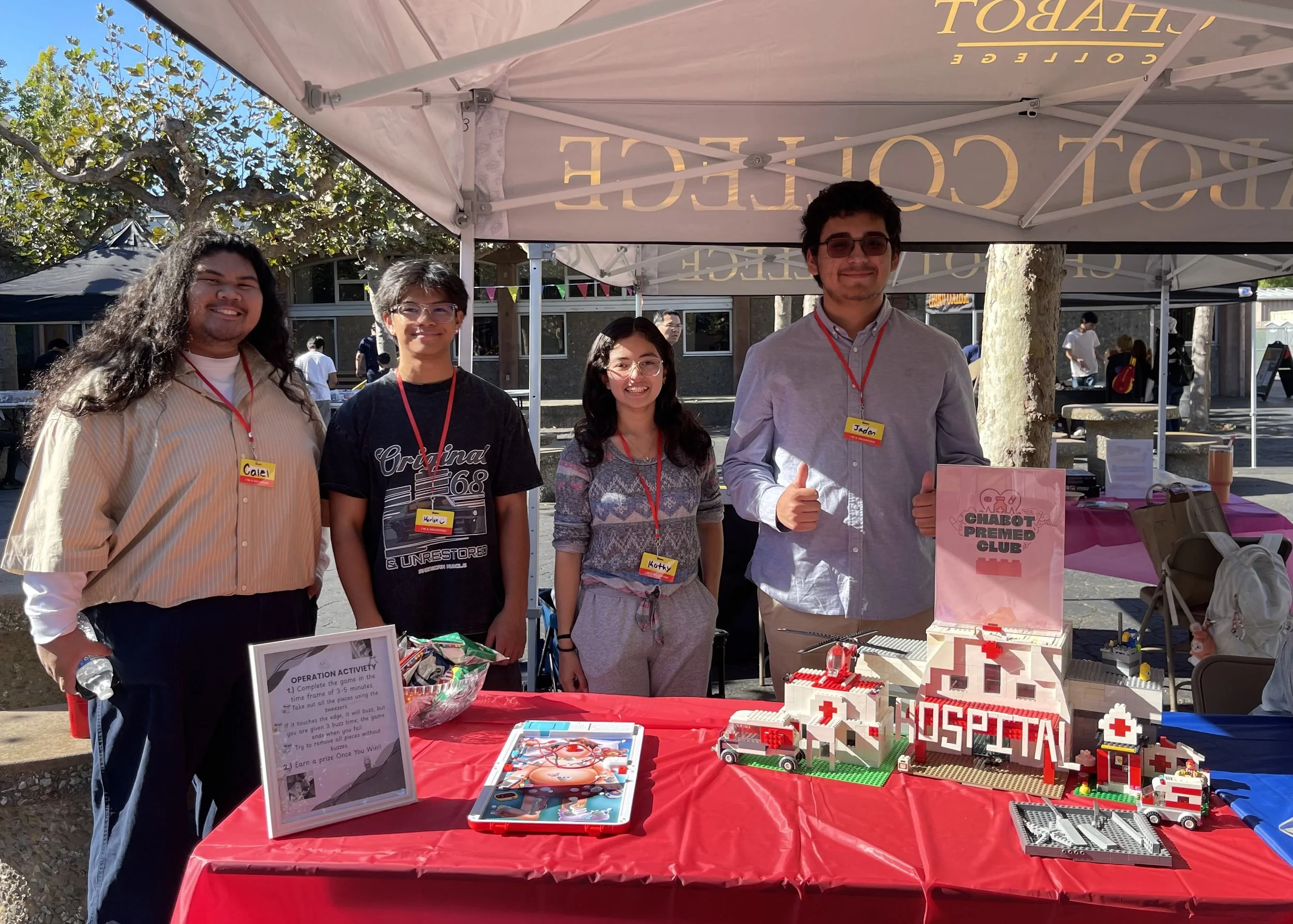 Five young people standing behind a table with LEGO hospital-themed displays at an outdoor event, under a white canopy with 'Chocolate' written on it, smiling and posing for the photo.