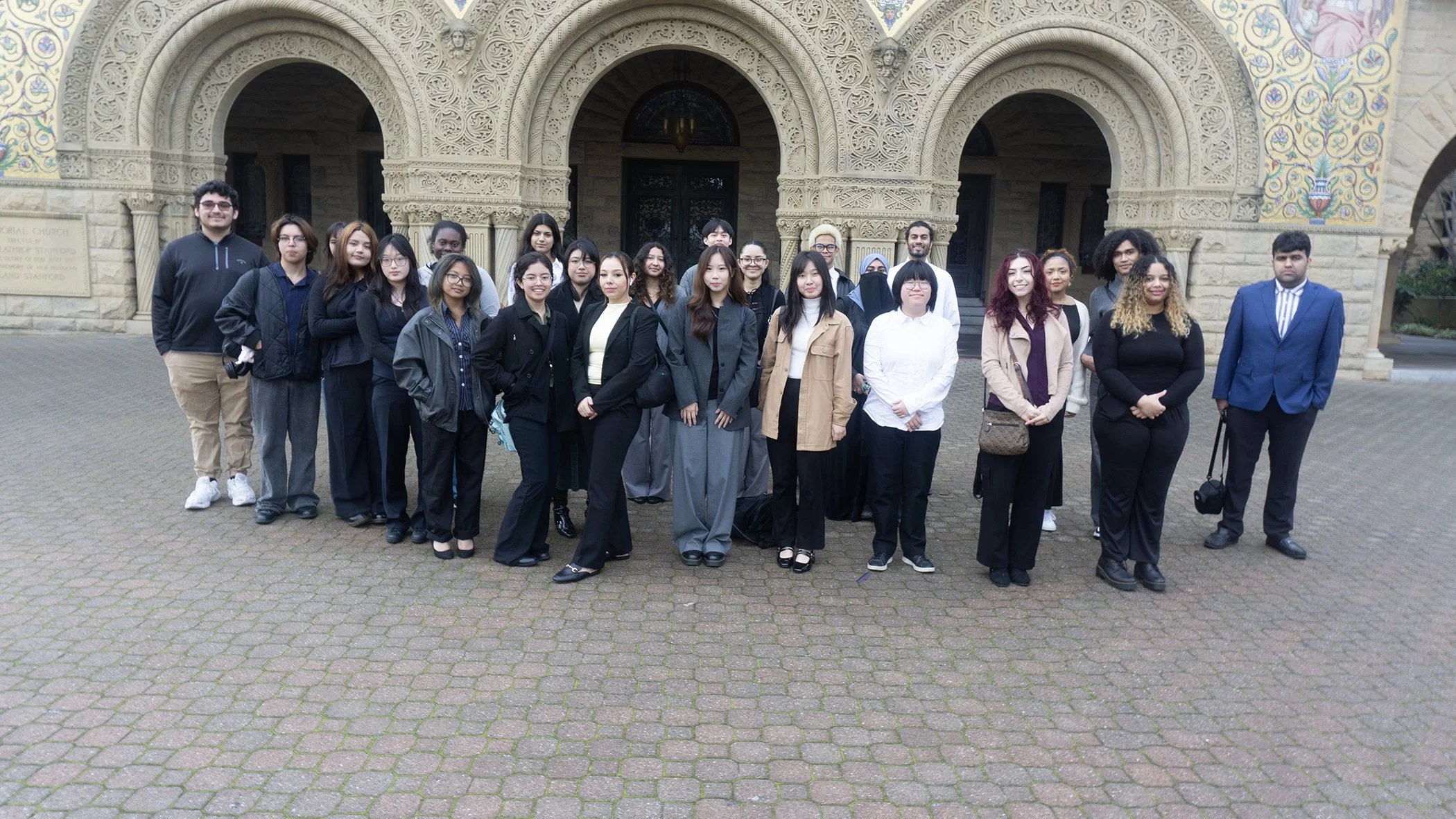 Group of diverse young adults standing in front of an ornate stone building with arches, some smiling at the camera.