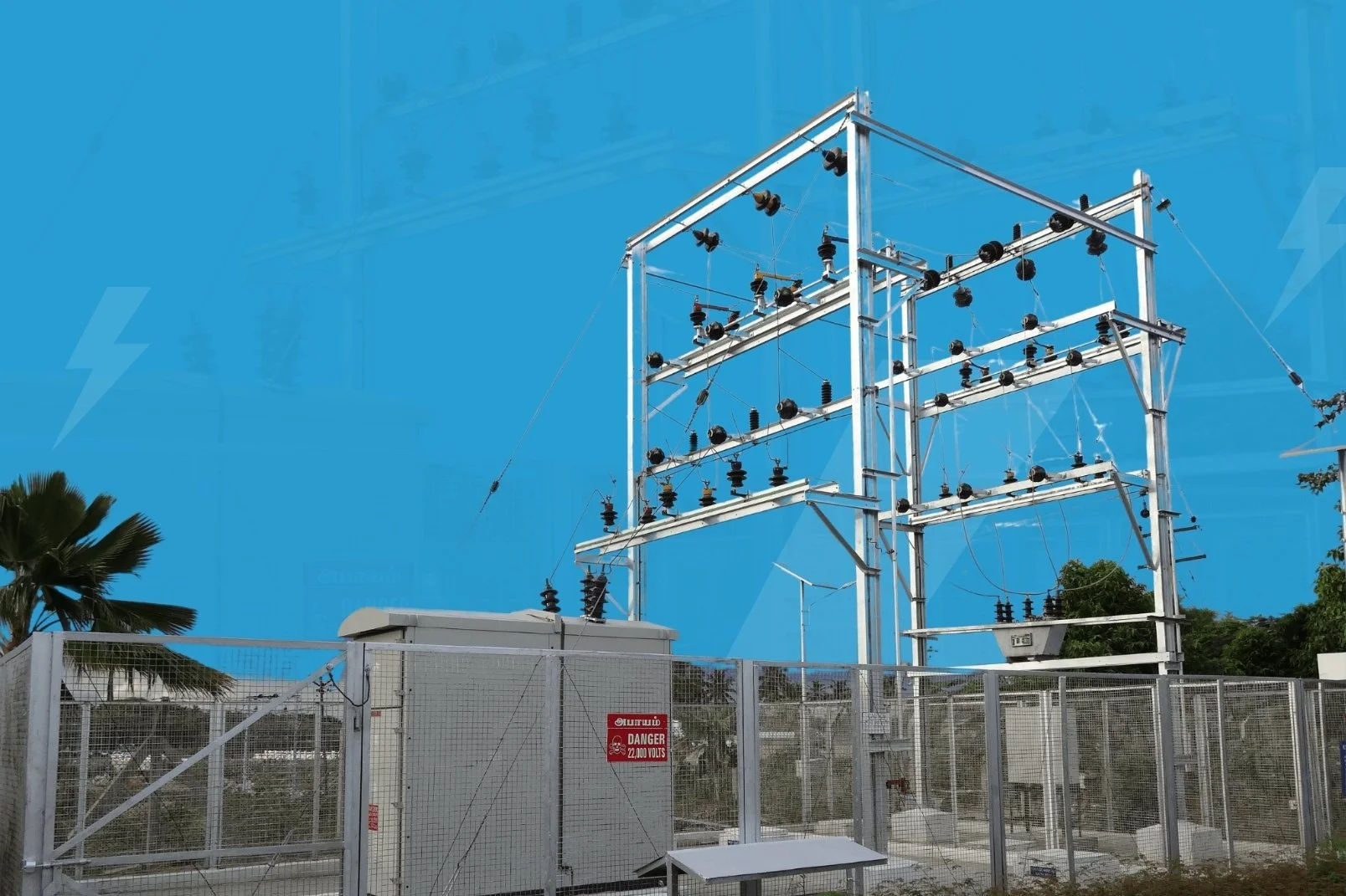 Electric power substation with metal structures, insulators, transformers, and wiring enclosed by a metal fence, with a bright blue sky and some trees in the background.