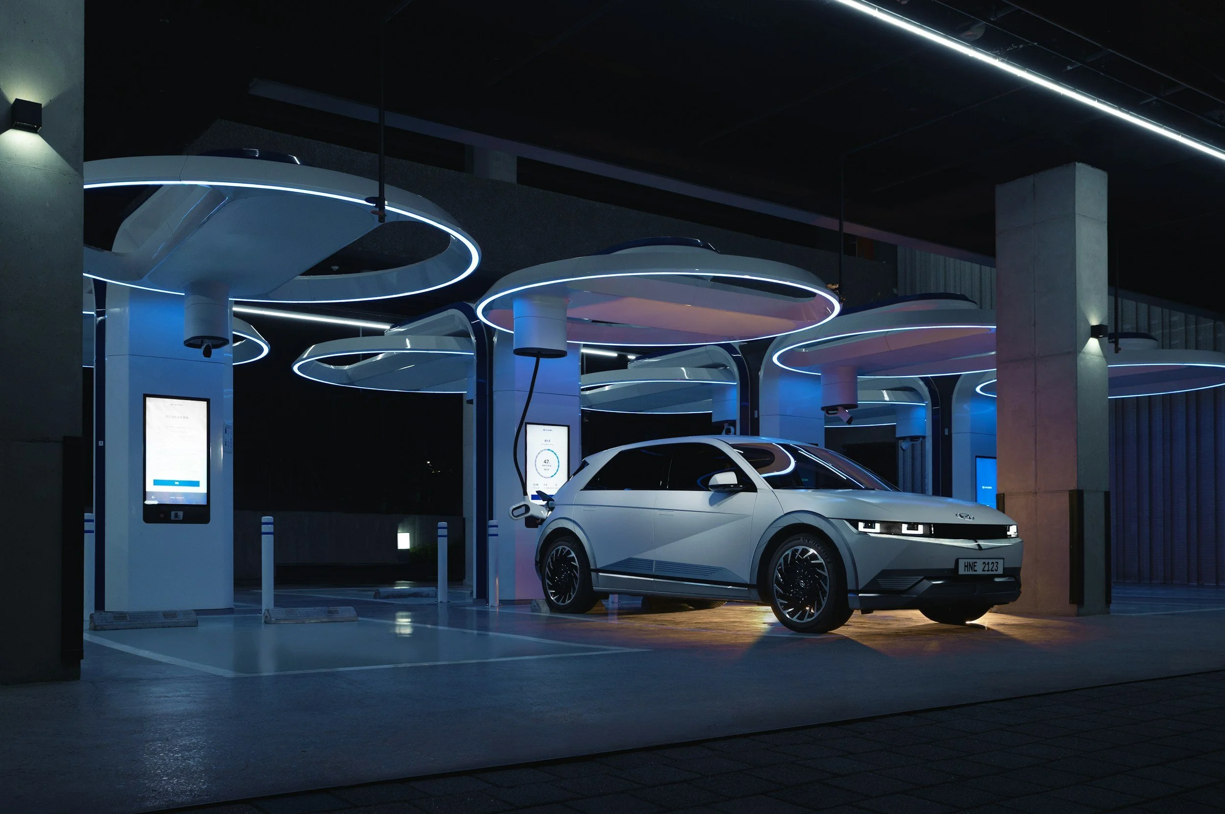 A white electric car parked at an advanced, futuristic EV charging station illuminated by blue LED lights at night.