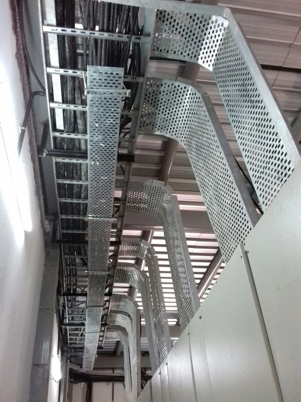 A row of metallic cable management trays installed in an electrical room, supporting and organizing black electrical cables along the ceiling.