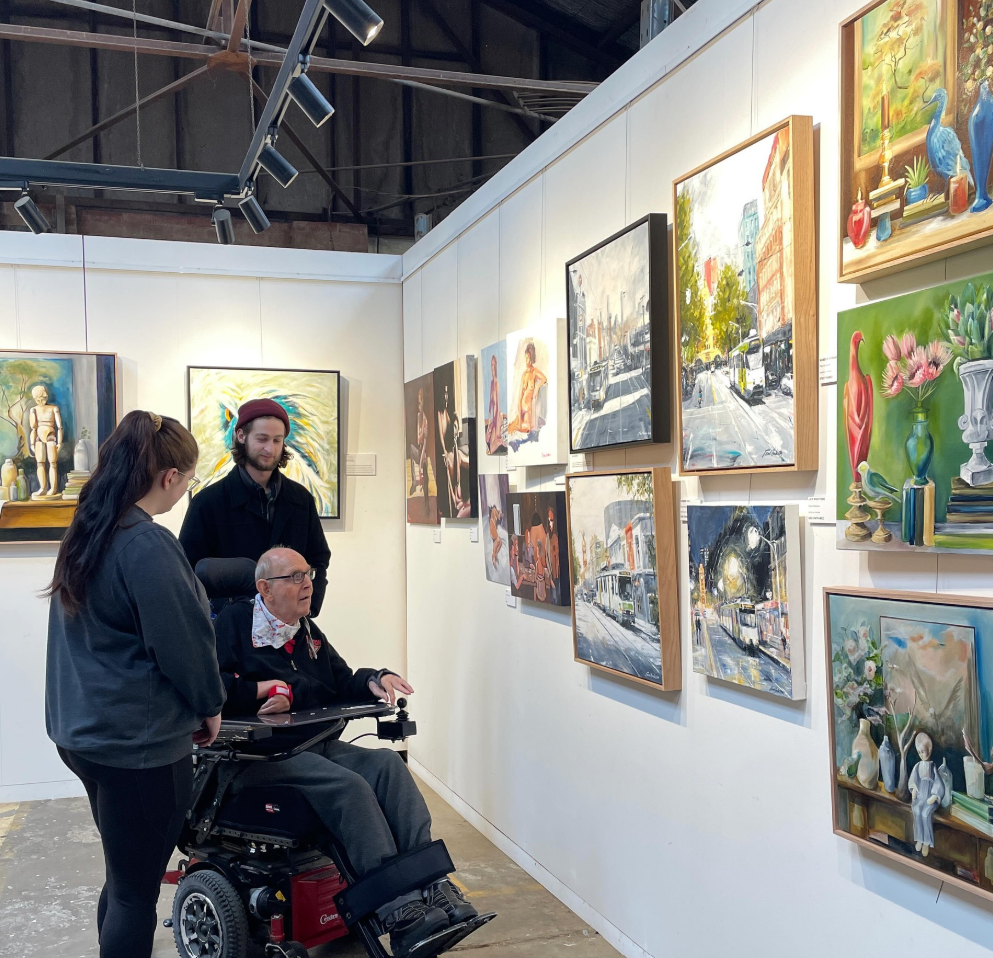 Three people looking at paintings in an art gallery, including a woman, a man with a beard, and an elderly man in a wheelchair, with various colorful paintings displayed on the white gallery walls.