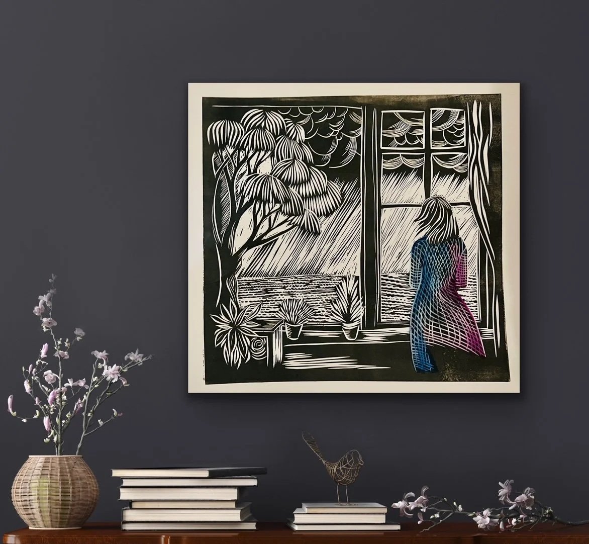 A black and white paper cutout artwork depicting a woman looking out a window at the sea, with plants and trees inside and outside, hanging on a dark wall. There is a bookshelf with books and a vase with flowers on a wooden shelf below the artwork.