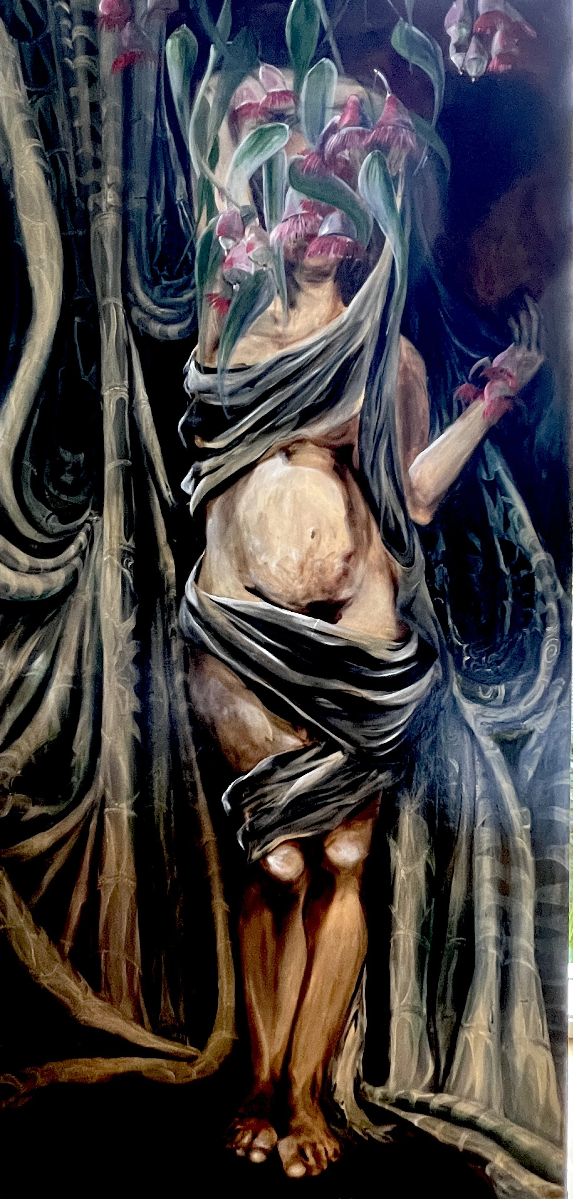 A surreal painting of a partially nude woman with a leafy headdress that covers her face. She is wrapped in dark cloths, surrounded by bamboo and textured backgrounds, creating an abstract, mysterious atmosphere.